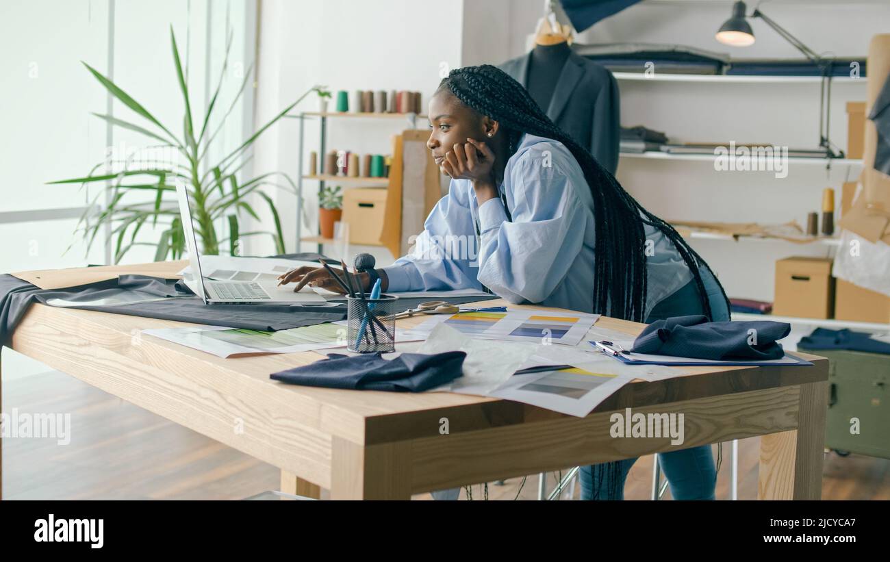 Un talentueux designer de mode afro-américain crée une nouvelle collection de mode tout en travaillant sur un ordinateur portable dans un studio Fashion Business Startup. Concept Banque D'Images