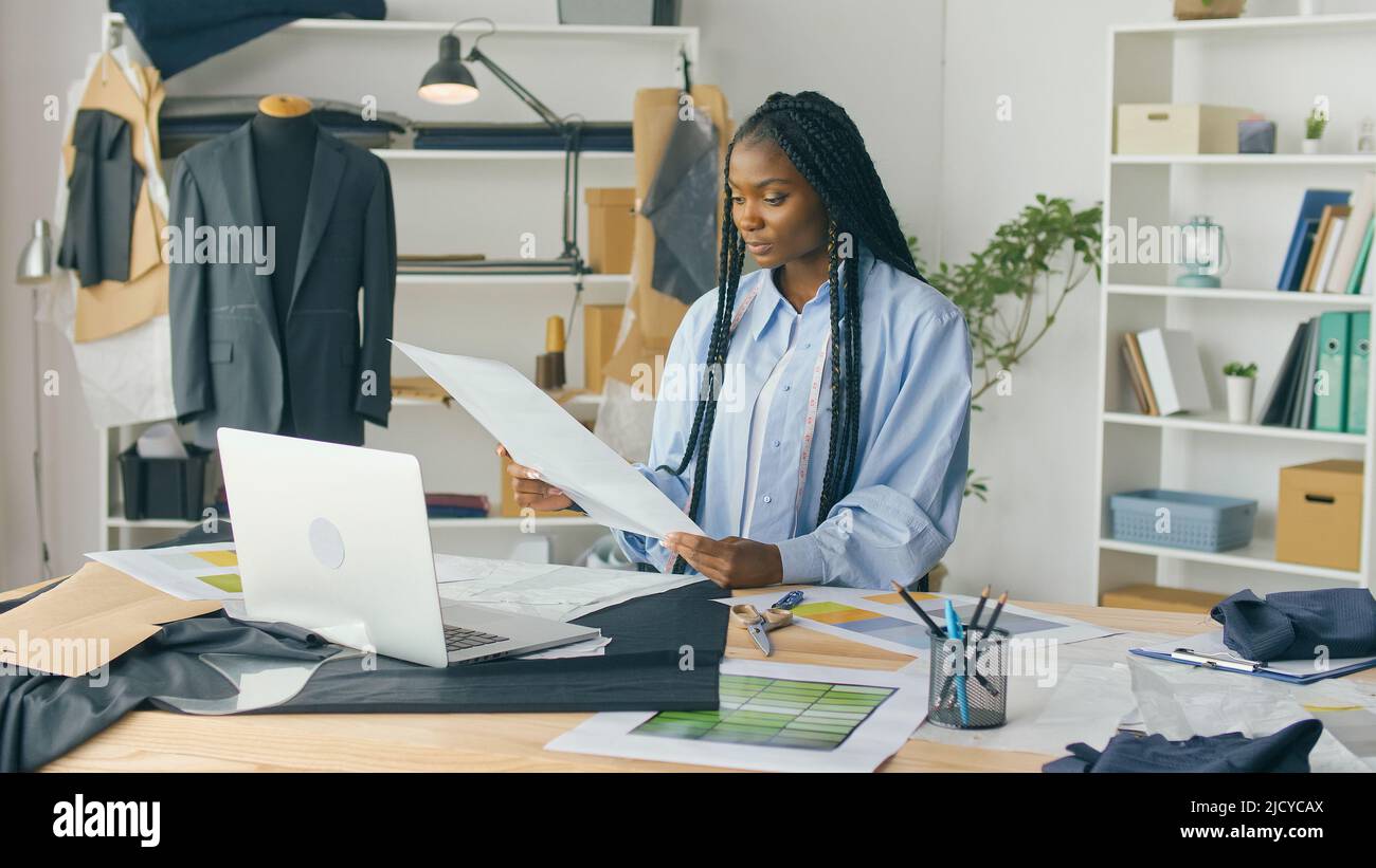 Un talentueux designer de mode afro-américain crée une nouvelle collection de mode tout en travaillant sur un ordinateur portable dans un studio Fashion Business Startup. Concept Banque D'Images