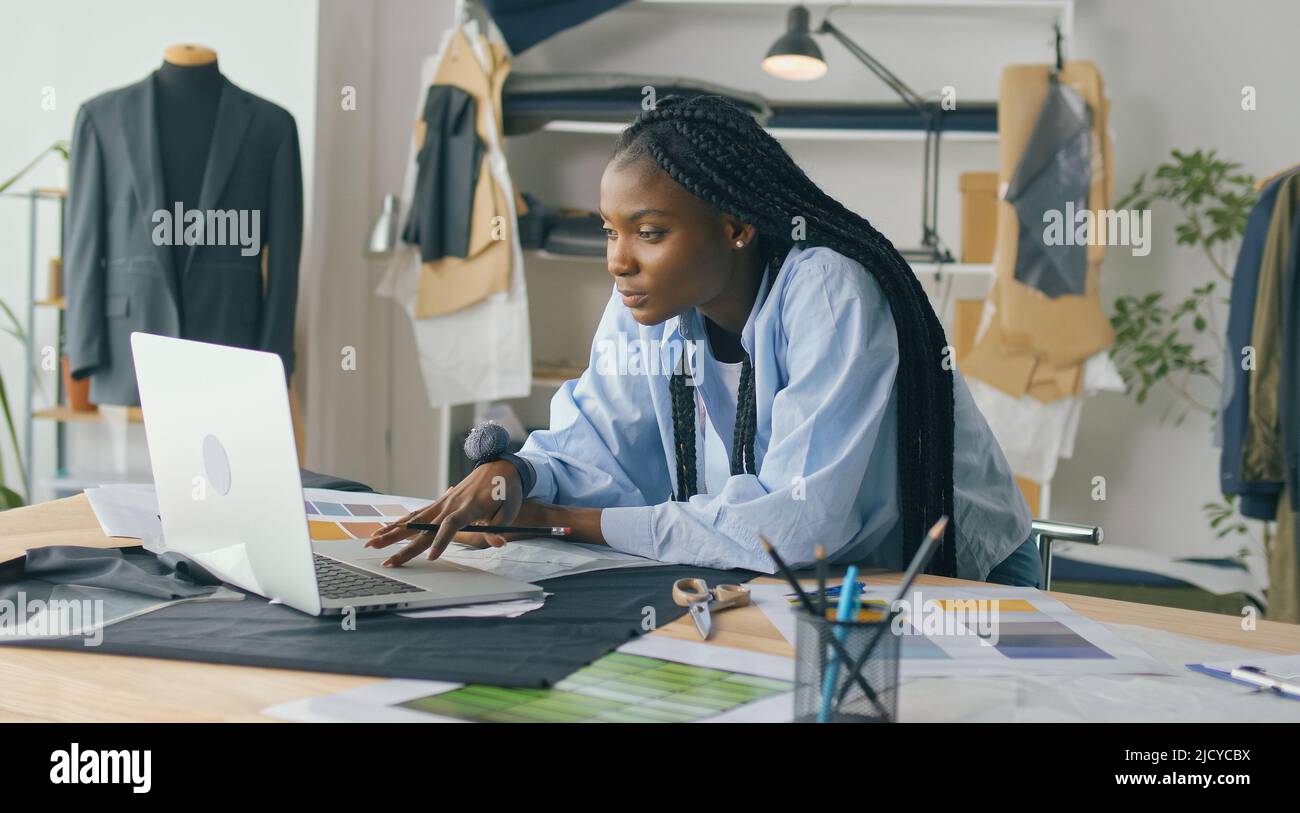 Un talentueux designer de mode afro-américain crée une nouvelle collection de mode tout en travaillant sur un ordinateur portable dans un studio Fashion Business Startup. Concept Banque D'Images