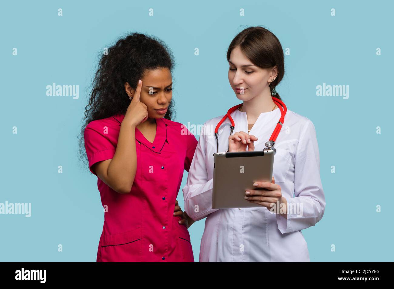 La femme caucasienne GP et la pensive afro-américaine femme médecin assistant stagiaire regarde la tablette numérique sur Blue Studio Wall. Équipe médicale multiethnique Banque D'Images