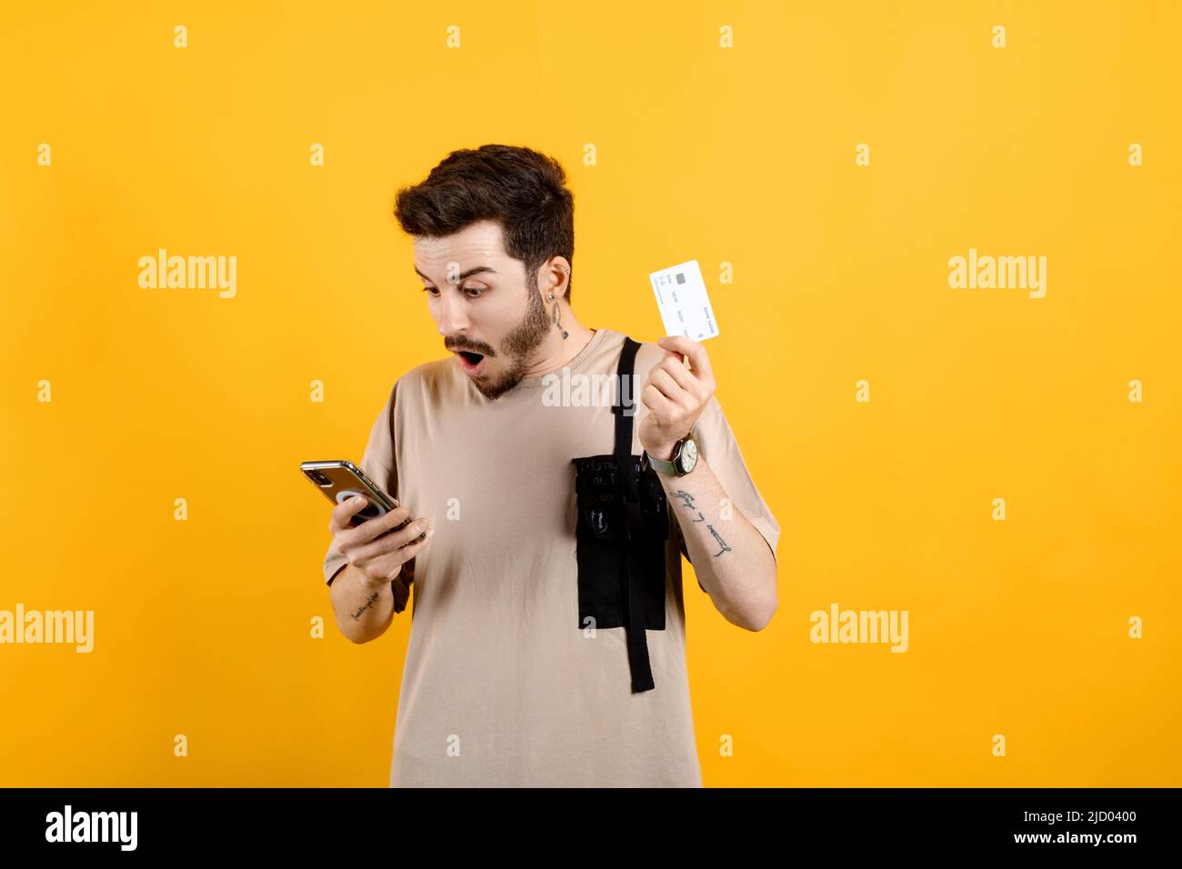 Homme caucasien portant le t-shirt beige posant isolé sur fond jaune tenant smartphone et carte de crédit choqué par la surprise et l'expression stupéfaite, Banque D'Images