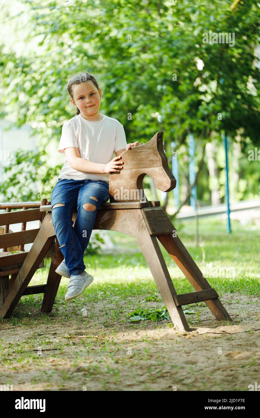 Jolie petite fille assise sur un cheval en bois dans une aire de jeux dans un parc verdoyant. Vacances d'été dans le camp, centre touristique. Marcher et jouer à l'extérieur, sport Banque D'Images