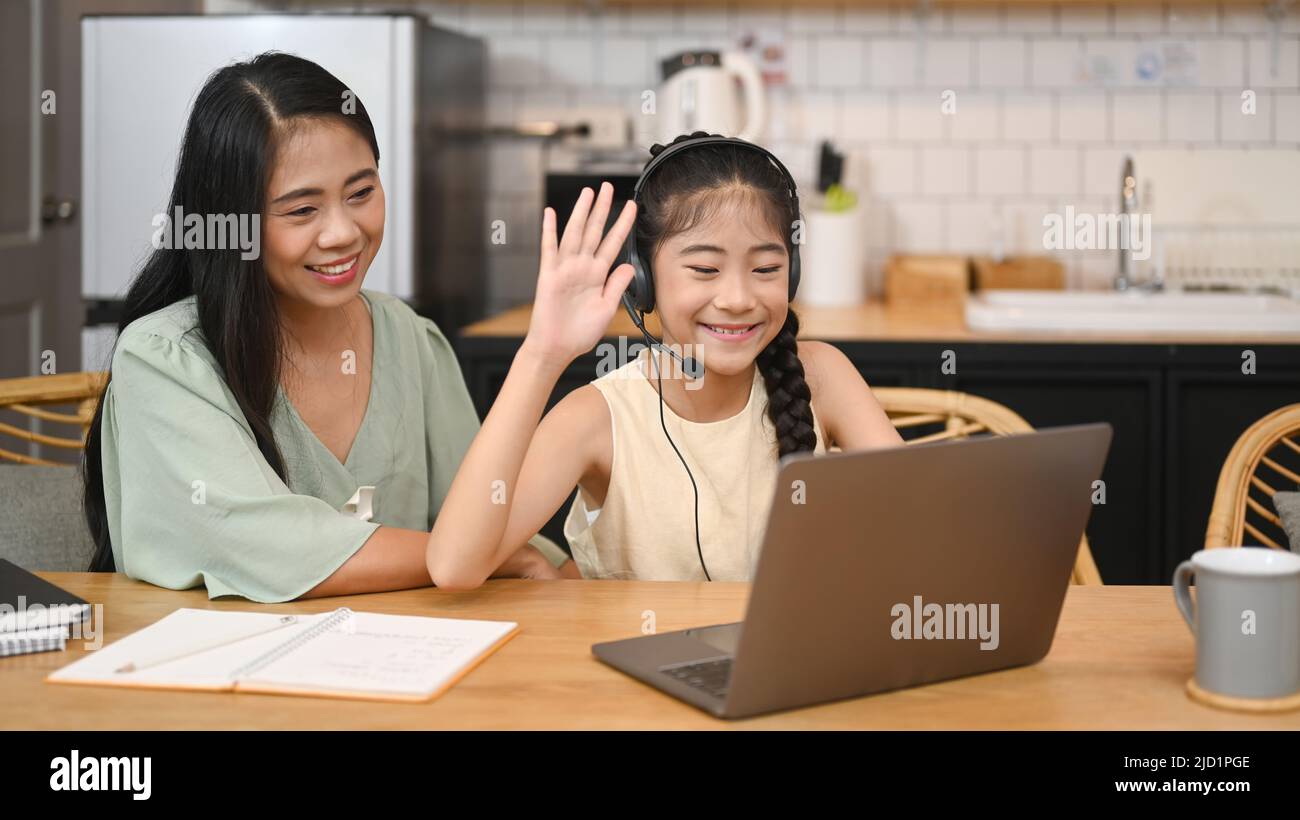 Une jeune fille asiatique gaie dans un casque qui parle à un professeur tout en étudiant en ligne via un ordinateur portable à la maison avec une maman heureuse assise à proximité et en donnant du soutien Banque D'Images