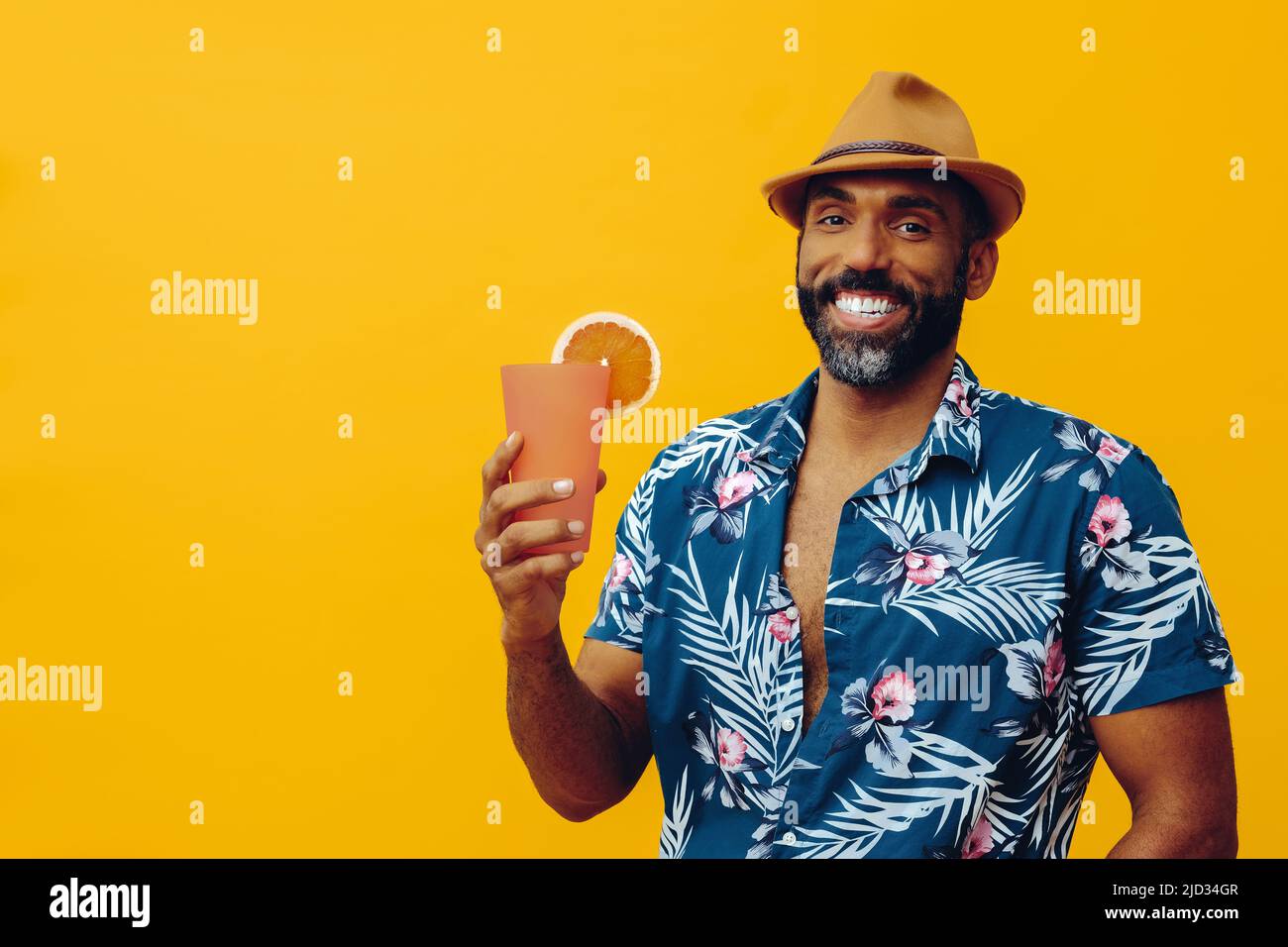 Beau barbu homme afro-américain adulte moyen portant une chemise hawaïenne et un chapeau souriant avec un cocktail de jus d'orange regardant le studio d'appareil photo Banque D'Images