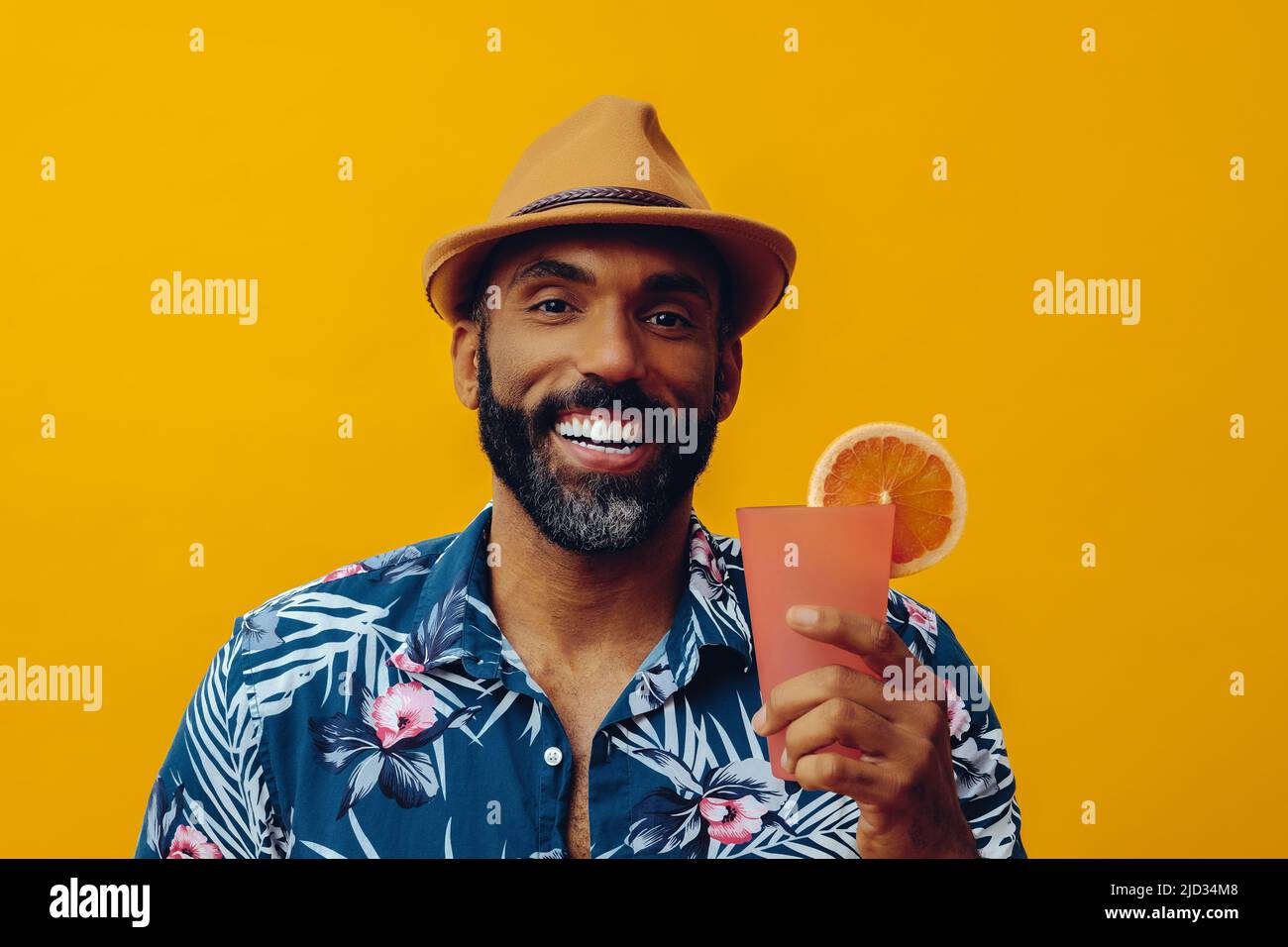 Homme afro-américain barbu adulte moyen portant une chemise hawaïenne et un chapeau avec un jus de cocktail orange souriant regardant l'appareil photo posé sur fond jaune studio Banque D'Images