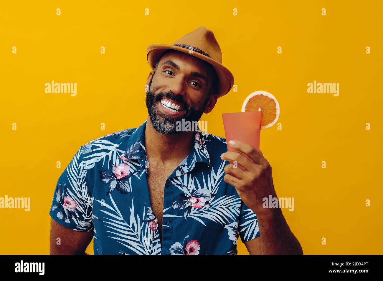 Joyeux homme américain africain de taille moyenne adulte portant une chemise hawaïenne et un chapeau souriant avec un cocktail de jus d'orange regardant le studio d'appareil photo Banque D'Images