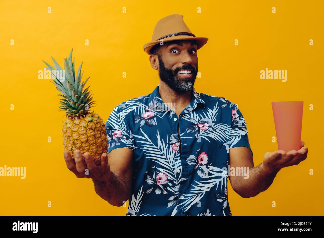 Homme afro-américain souriant, adulte moyen, regardant l'appareil photo portant une chemise hawaïenne et un chapeau équilibrant l'ananas et le verre avec les mains au studio Sides Banque D'Images