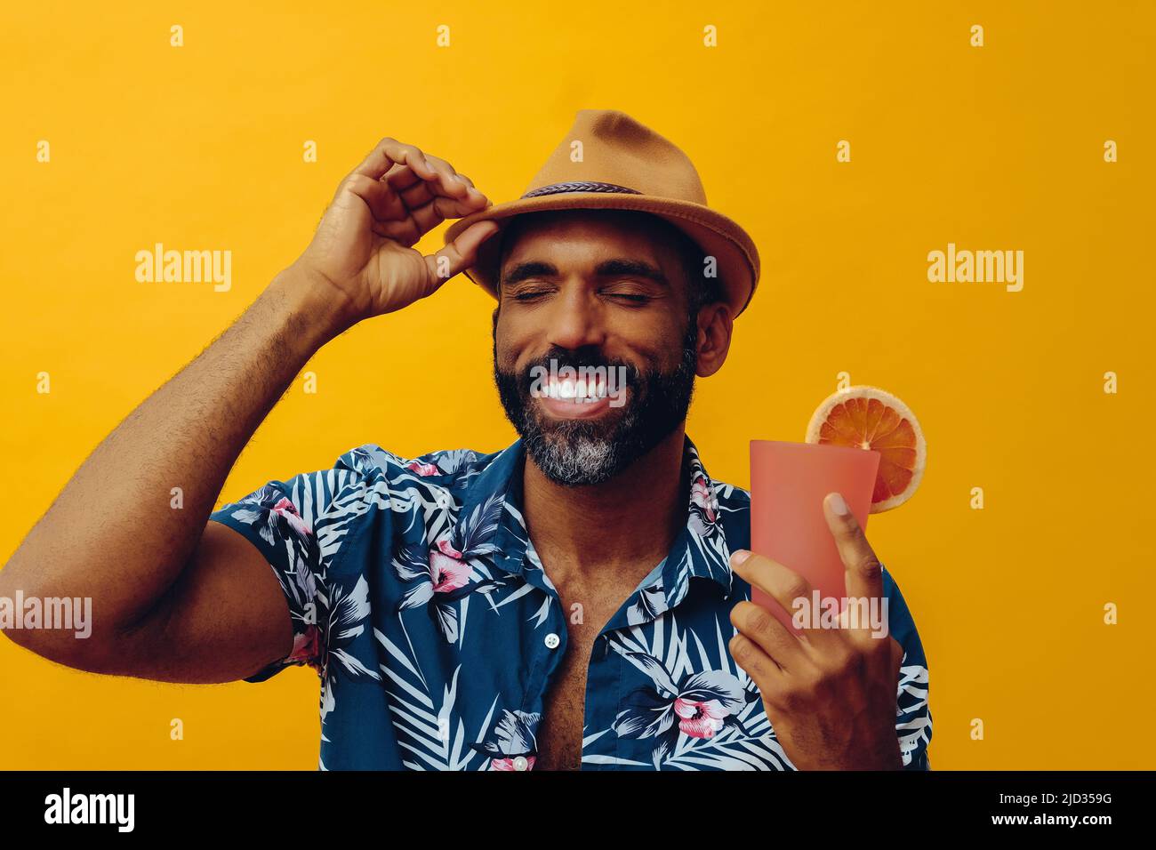 Homme afro-américain barbu adulte de taille moyenne portant une chemise hawaïenne et un chapeau à la main avec du jus de cocktail orange souriant avec les yeux fermés studio Banque D'Images