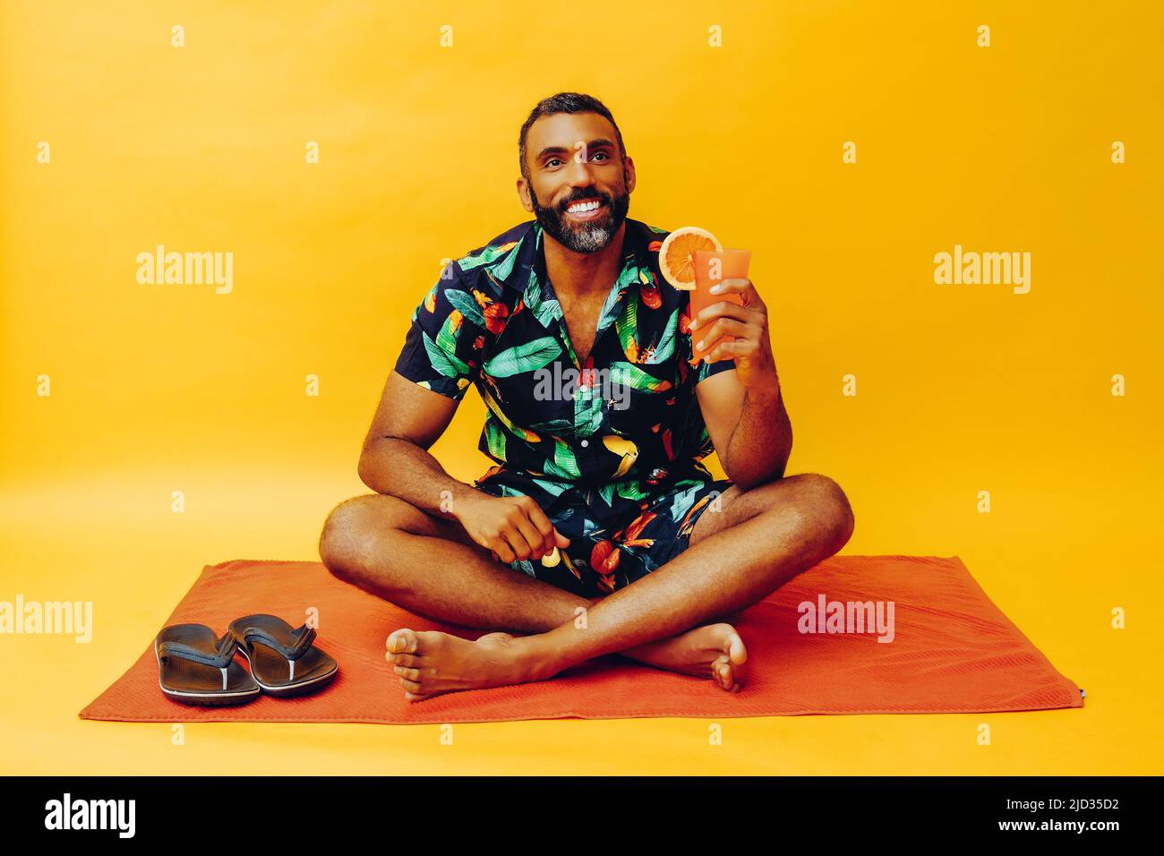 beau barbu homme afro-américain moyen adulte souriant pendant les vacances assis sur une serviette orange, tenant du jus d'orange regardant la prise de vue en studio Banque D'Images