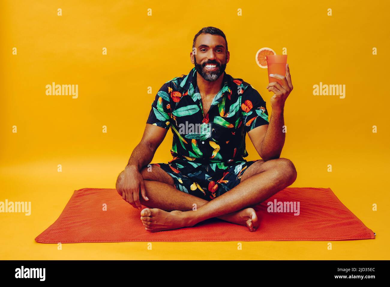 beau barbu homme afro-américain moyen adulte souriant pendant les vacances assis sur une serviette orange, tenant du jus d'orange regardant la prise de vue en studio Banque D'Images