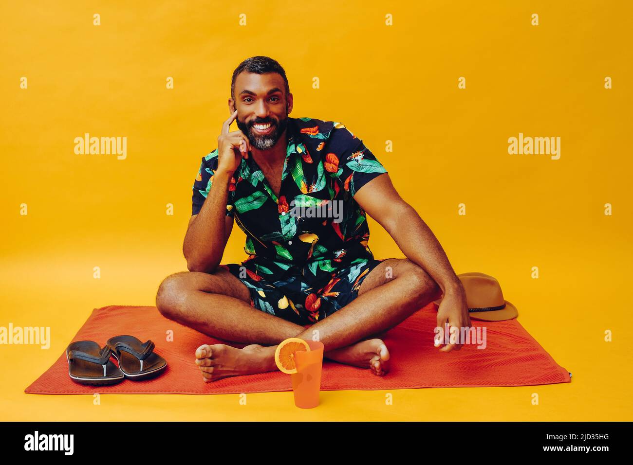 beau barbu homme afro-américain moyen adulte souriant pendant les vacances assis sur une serviette orange, tenant du jus d'orange regardant la prise de vue en studio Banque D'Images