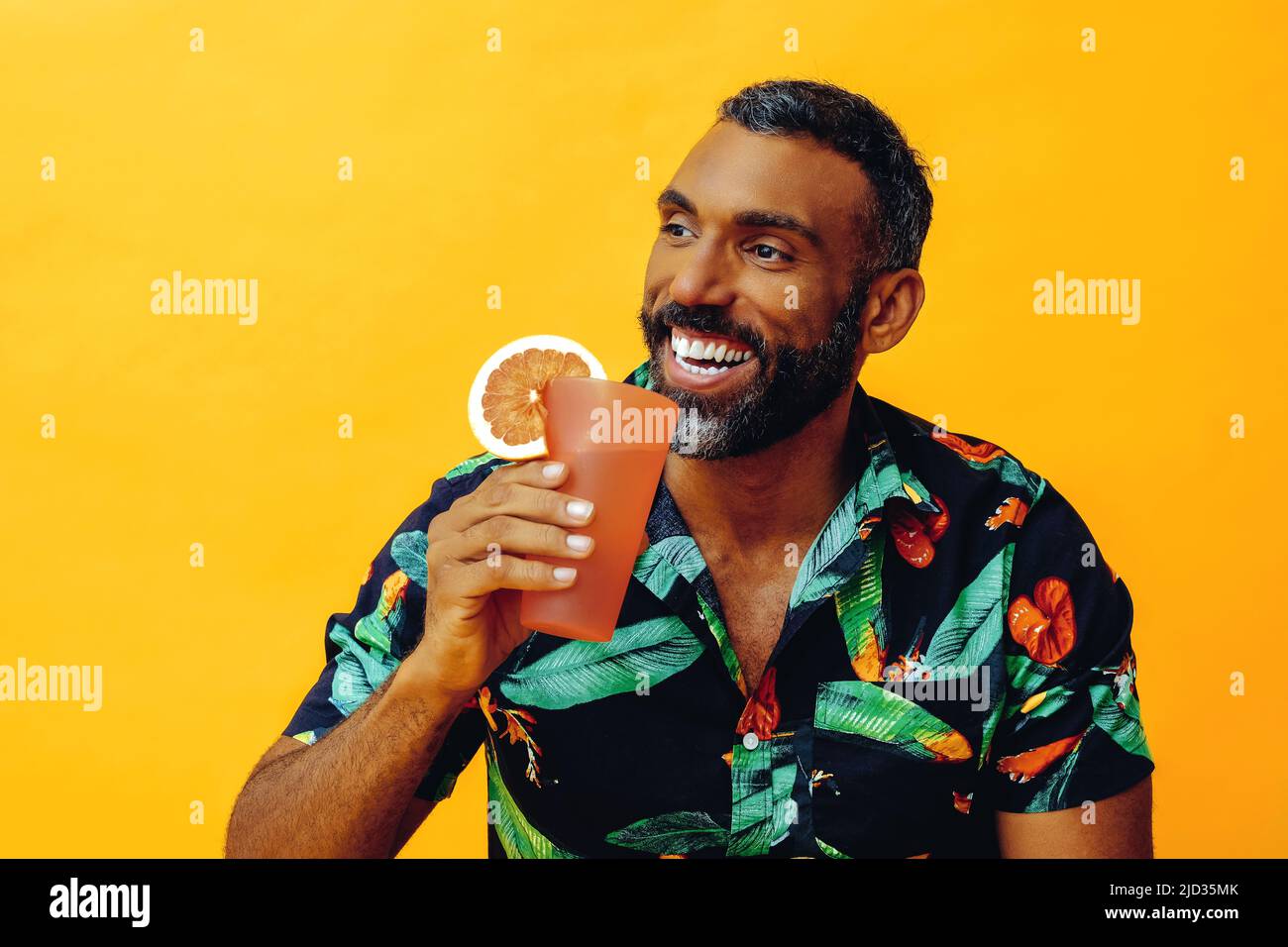 beau barbu adulte moyen homme afro-américain souriant en vacances assis sur une chaise buvant un cocktail de jus d'orange en studio Banque D'Images