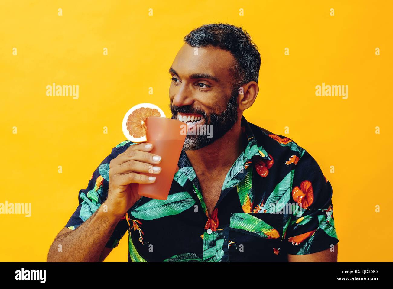 beau barbu adulte moyen homme afro-américain souriant en vacances assis sur une chaise buvant un cocktail de jus d'orange en studio Banque D'Images