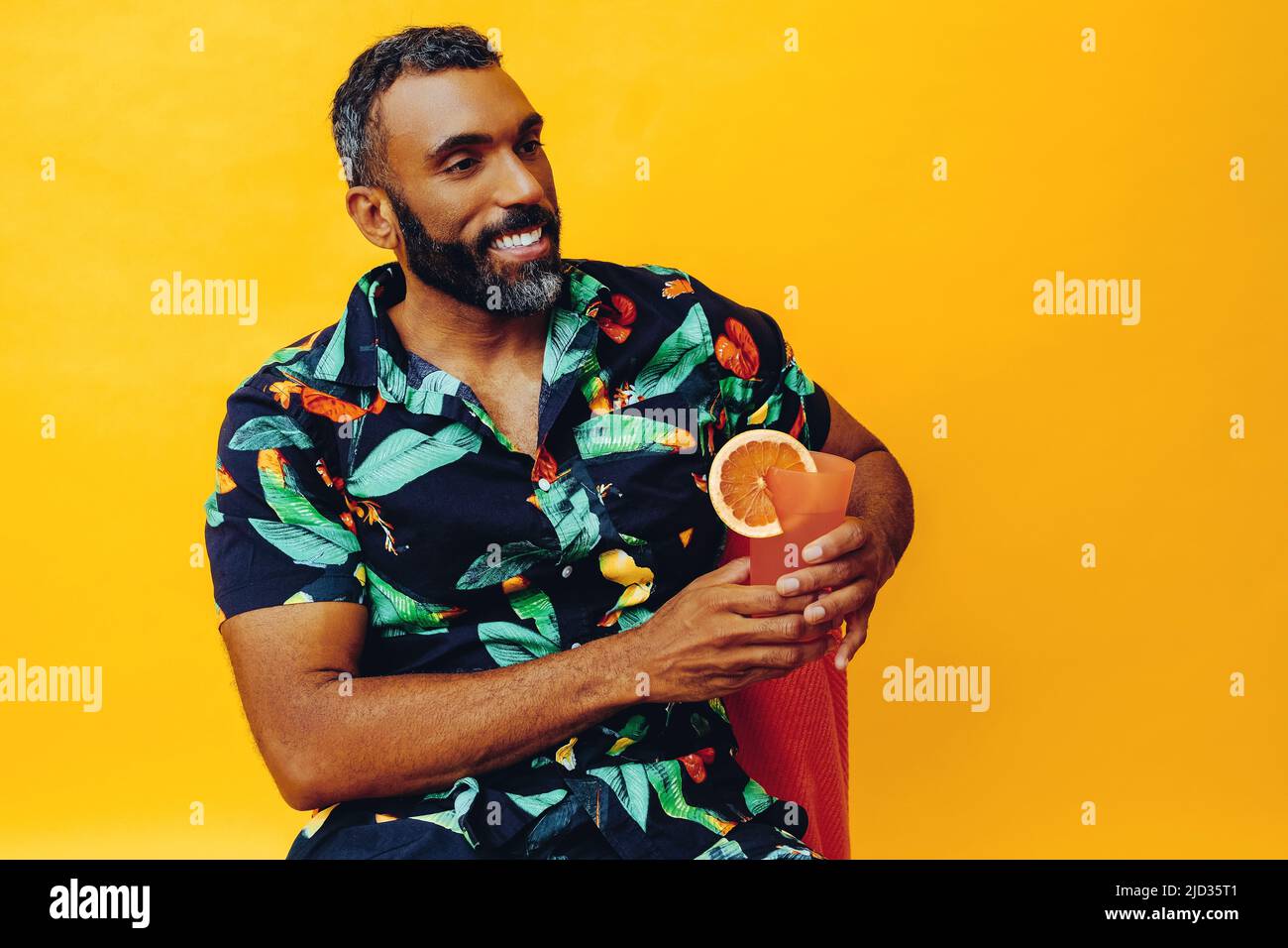 beau barbu homme afro-américain moyen adulte souriant pendant les vacances assis sur une serviette orange, tenant le jus d'orange regardant loin prise de vue studio Banque D'Images