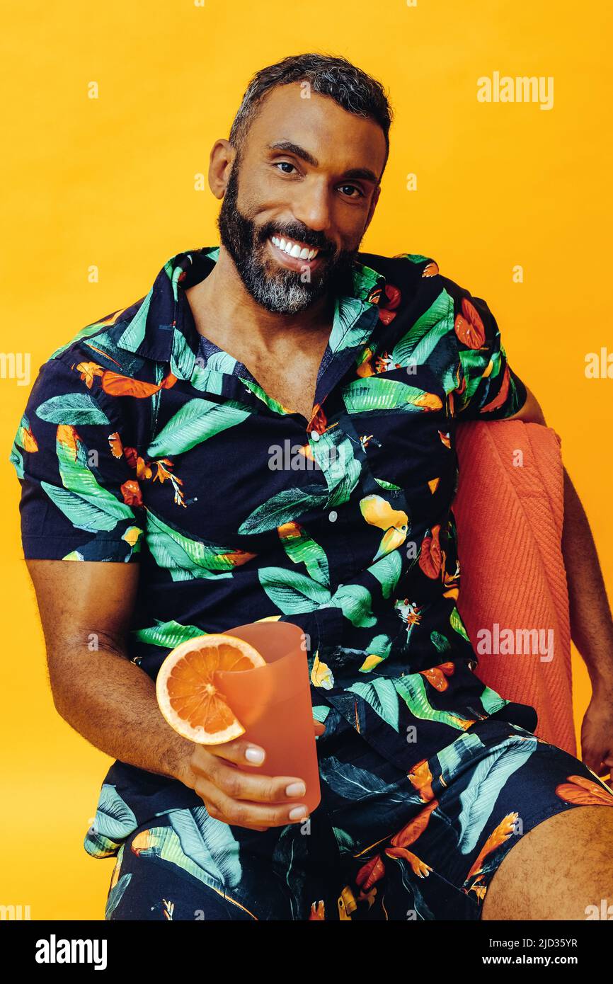 beau barbu homme afro-américain moyen adulte souriant pendant les vacances assis sur une serviette orange, tenant du jus d'orange regardant la prise de vue en studio Banque D'Images