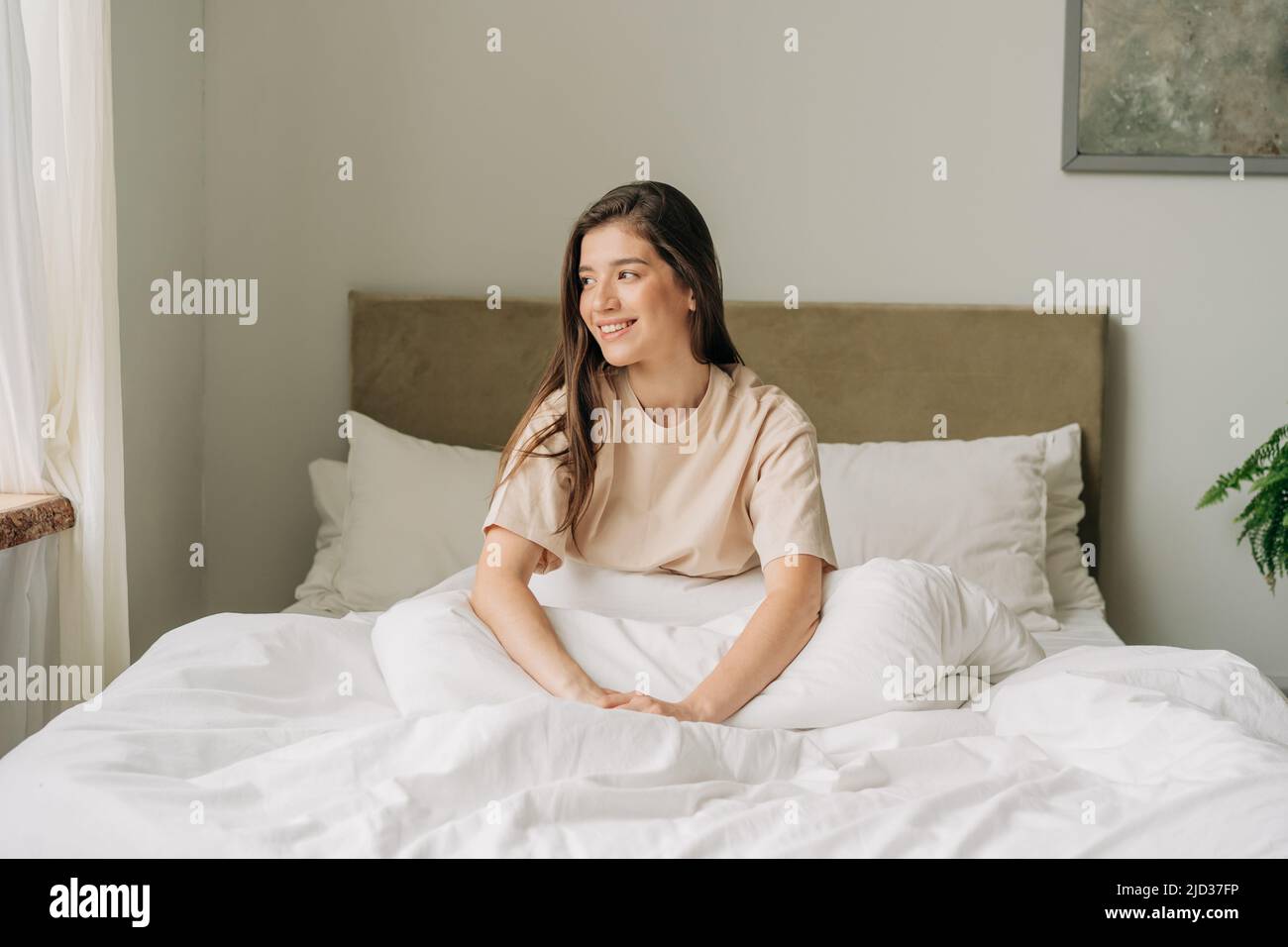 Jeune femme brune souriante reposée assise sur le lit dans une chambre lumineuse et confortable tôt le matin Banque D'Images