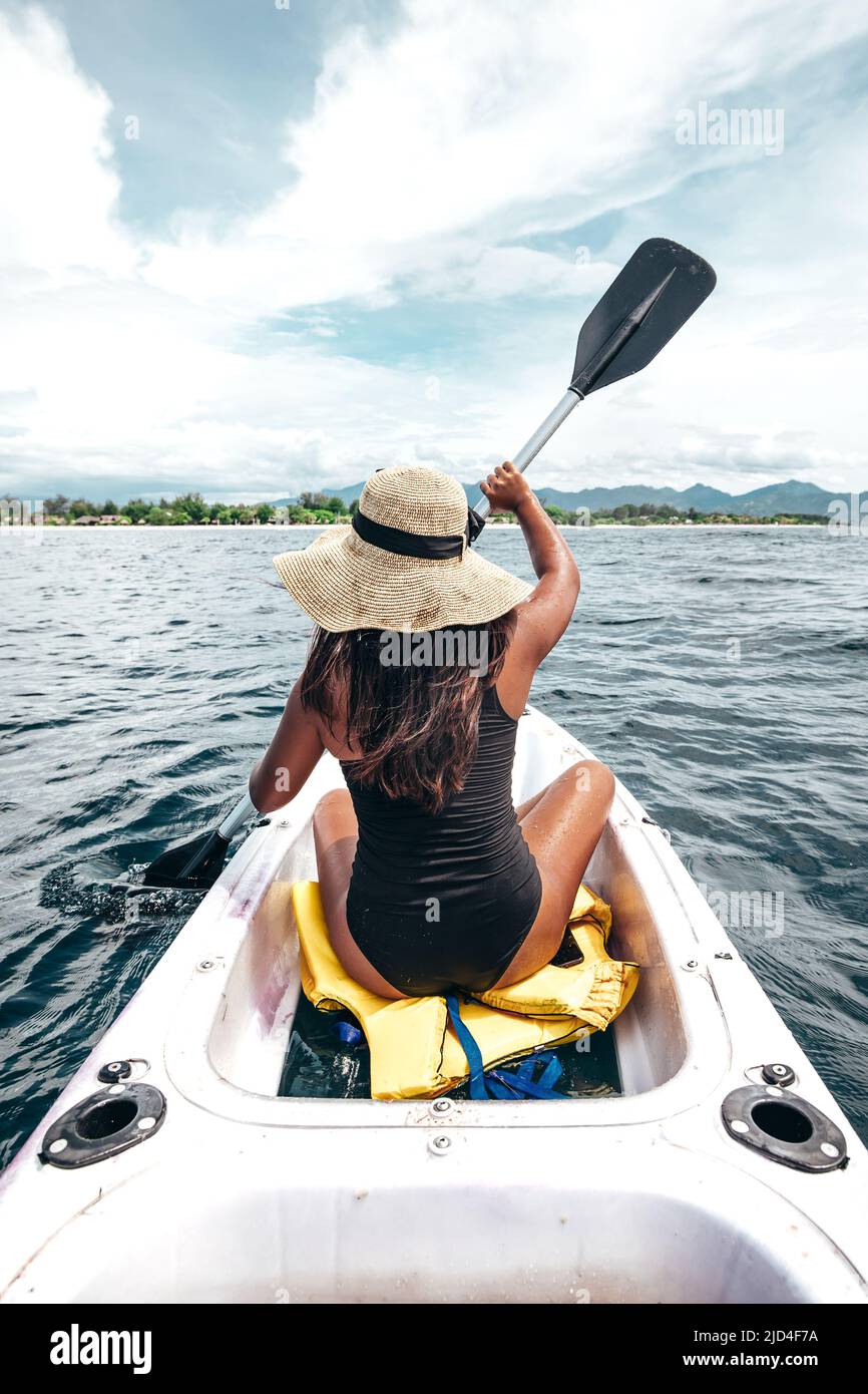 Jeune femme indonésienne en maillot de bain noir et chapeau pagayant en kayak blanc Banque D'Images