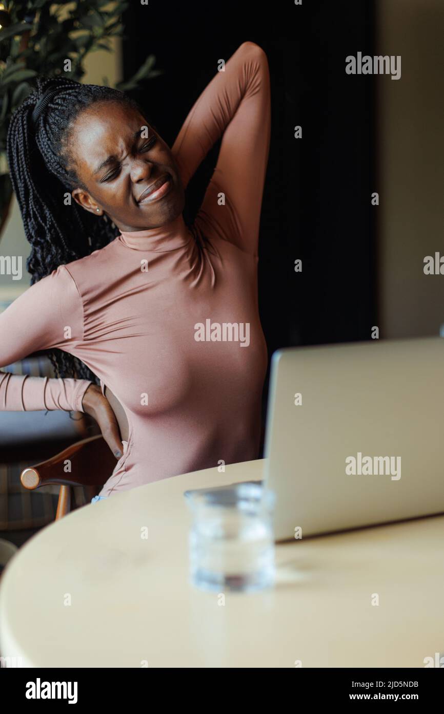 Afro-américaine dans un joli col de cygne qui s'étire avec les yeux fermés assis sur une table et qui travaille sur un ordinateur portable dans un café. Fille noire travaillant Banque D'Images