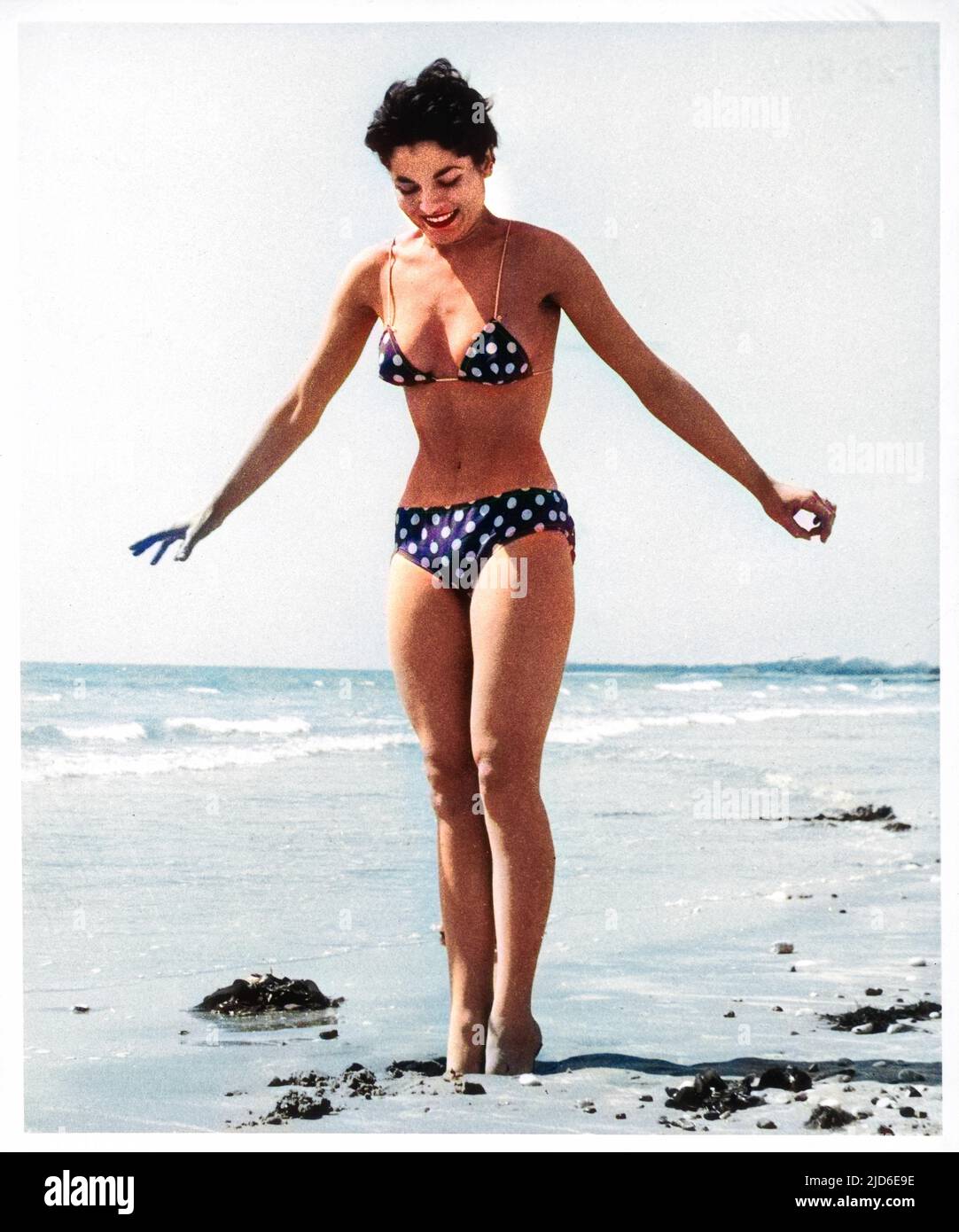 Une jeune femme athlétique, portant un bikini à pois et à fines bretelles spaghetti, s'enfonce dans le sable humide sur une plage. Version colorisée de : 10139497 Date: 1950s Banque D'Images