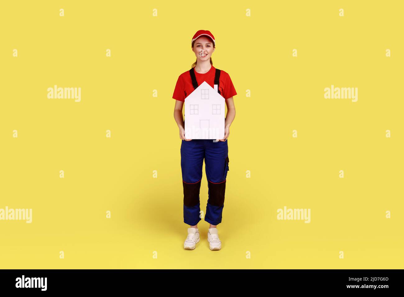 Portrait complet de la femme travailleuse motionnaire offrant de l'aide dans la réinstallation, les services de réparation à domicile et l'entretien, le port de combinaisons et de casquette rouge. Studio d'intérieur isolé sur fond jaune. Banque D'Images