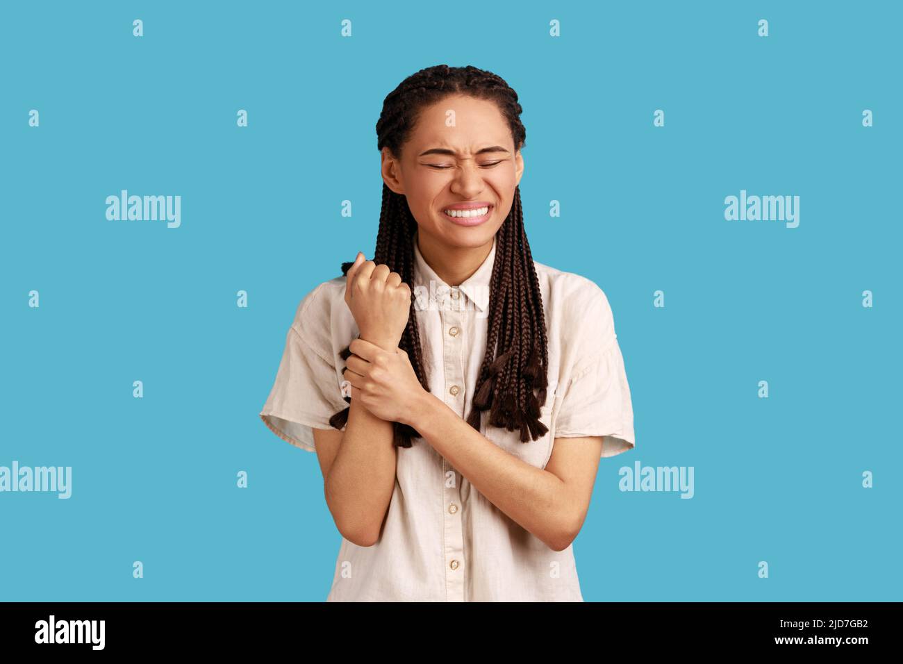 Contrarié mécontent femme avec des dreadlocks touche la main blessée, montre la zone douloureuse problématique sur le poignet, souffre de douleur terrible, portant une chemise blanche. Studio d'intérieur isolé sur fond bleu. Banque D'Images