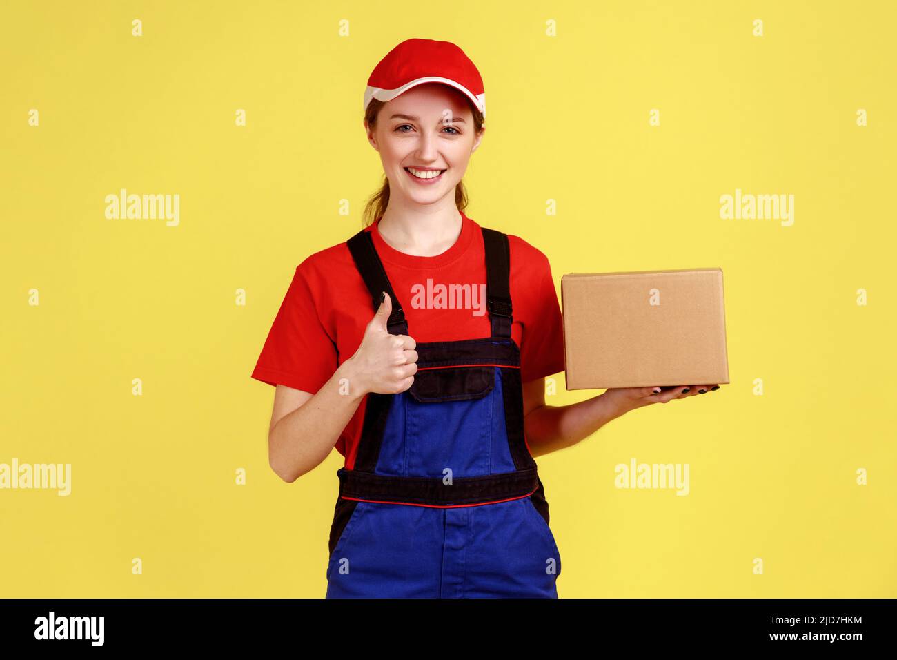 Portrait d'une femme de messagerie ravie debout avec une boîte en carton,  montrant le pouce vers le haut, comme le service de livraison, portant une  combinaison et une casquette rouge. Studio d'intérieur