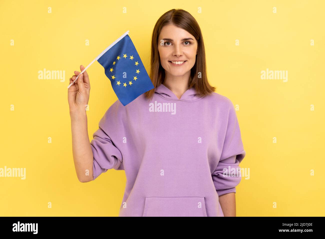 Femme patriotique aux cheveux foncés tenant le drapeau de l'ue à la main regardant l'appareil photo avec un sourire goûtant, patriotisme, célébration du jour du drapeau, portant le sweat à capuche violet. Studio d'intérieur isolé sur fond jaune. Banque D'Images