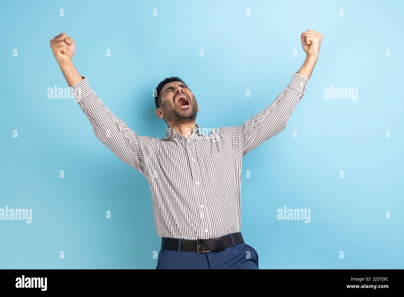 Portrait d'un homme d'affaires doué exprimant un geste gagnant avec des poings relevés et criant, célébrant la victoire, portant une chemise rayée. Studio d'intérieur isolé sur fond bleu. Banque D'Images Portrait d'un homme d'affaires doué exprimant un geste gagnant avec des poings relevés et criant, célébrant la victoire, portant une chemise rayée. Studio d'intérieur isolé sur fond bleu. Banque D'Images