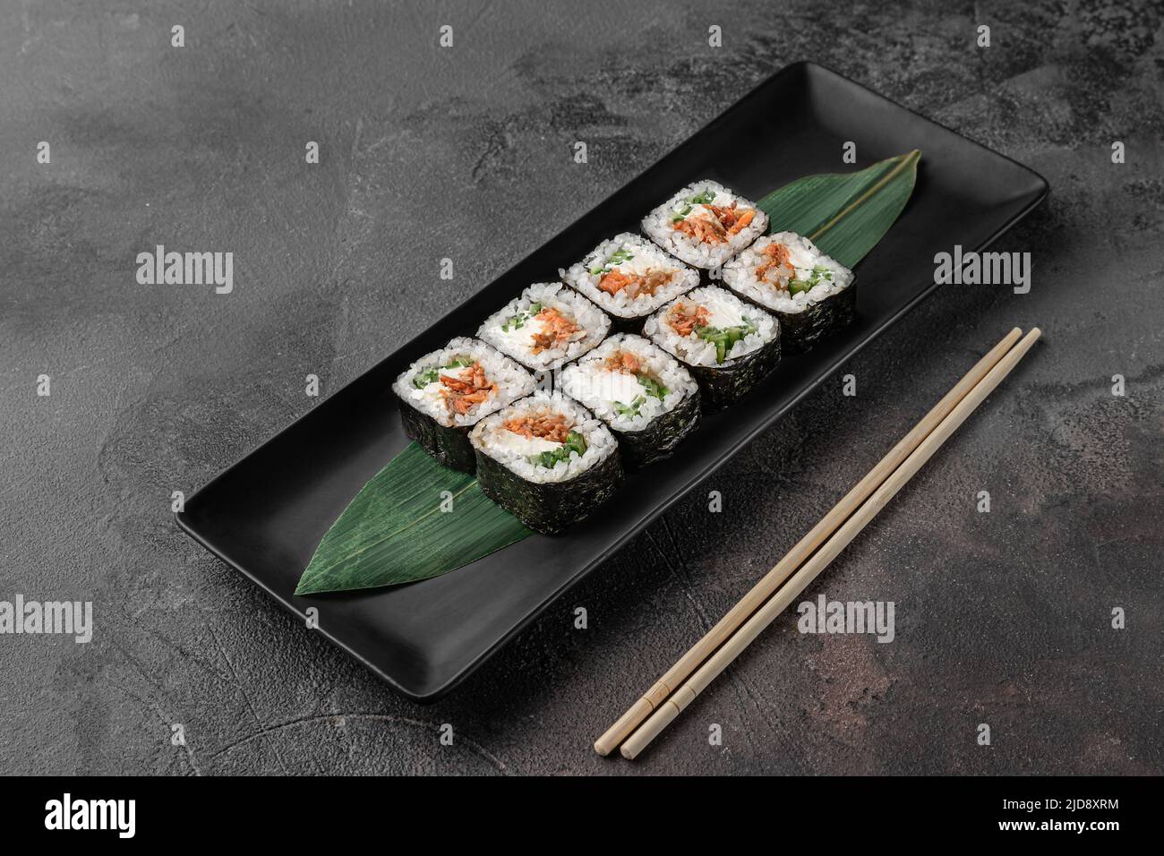 ensemble de rouleaux de maki carrés avec saumon frit, fromage caillé et feuille de bambou vert dans une plaque en céramique noire avec baguette sur un fond texturé gris foncé Banque D'Images