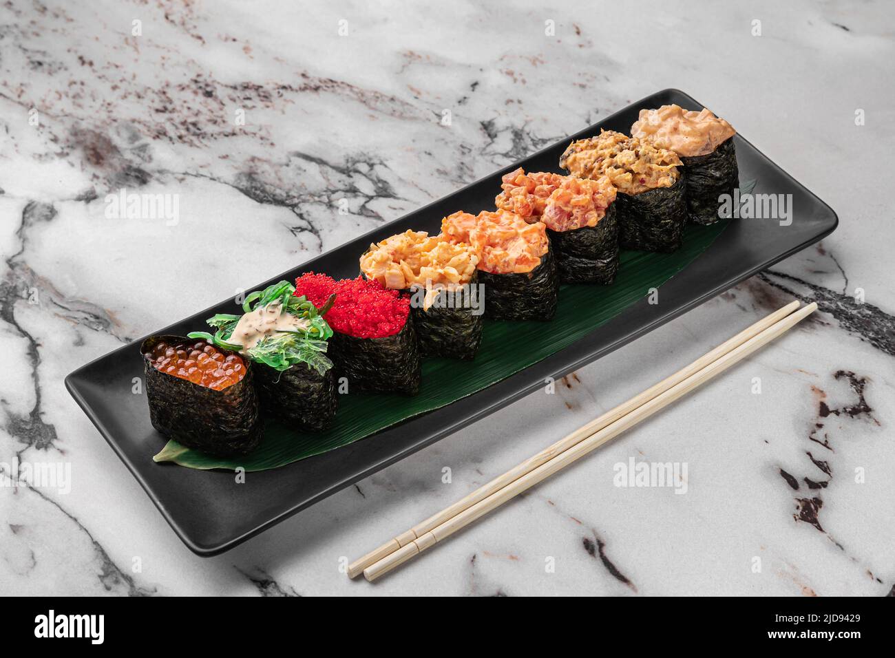 ensemble de gunkan avec saumon, salade de chuka, œufs de poisson volantes, thon et feuille de bambou vert dans une assiette en céramique noire avec baguette sur une texture blanche brillante Banque D'Images