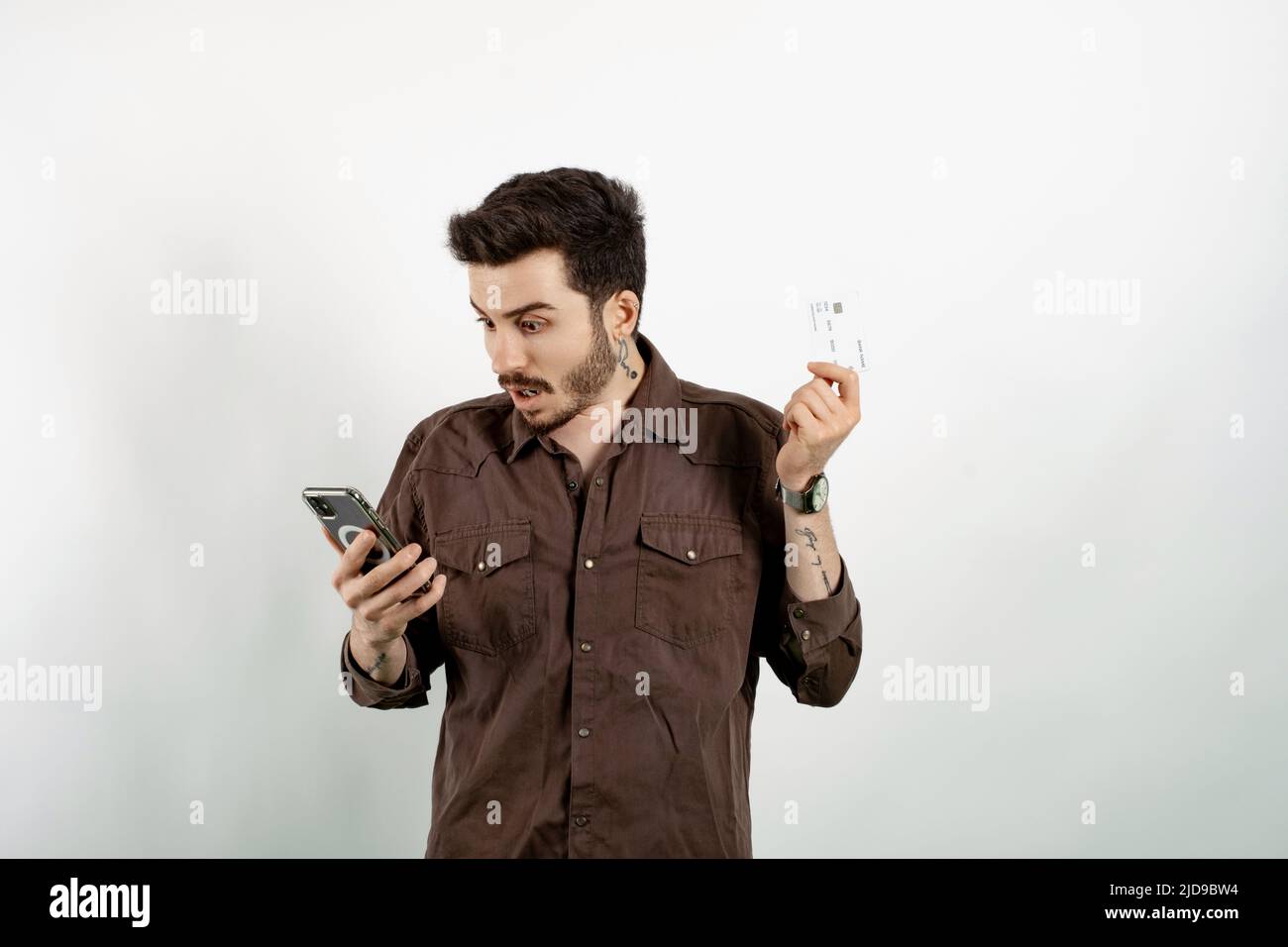 Homme caucasien portant une chemise marron posant isolé sur fond blanc tenant smartphone et carte de crédit choqué par surprise et expression stupéfait Banque D'Images