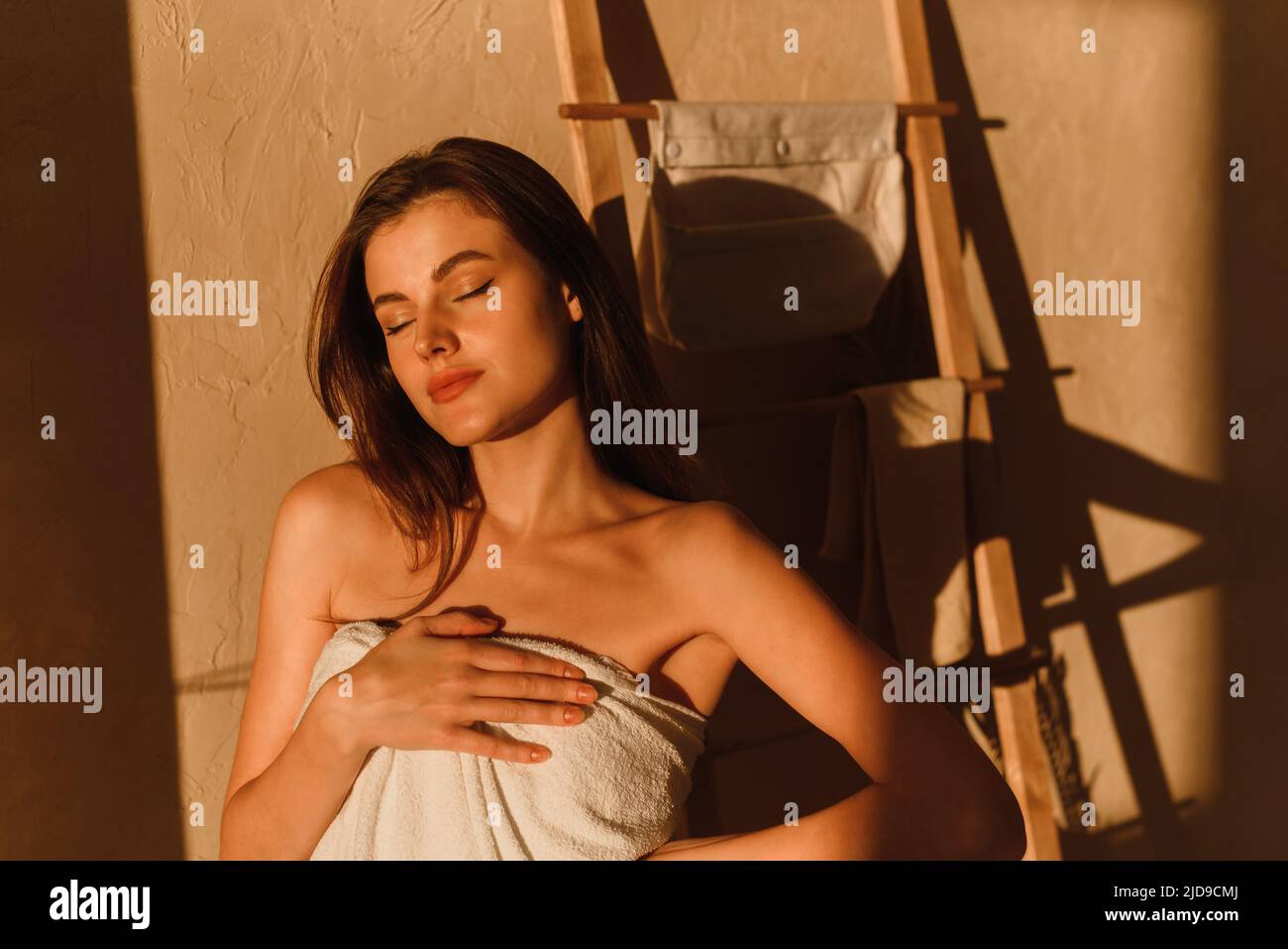 Une jeune femme profite du soleil. Une belle mannequin enveloppée d'une serviette blanche se tient dans la salle de bains après une douche. Ombres tropicales Banque D'Images
