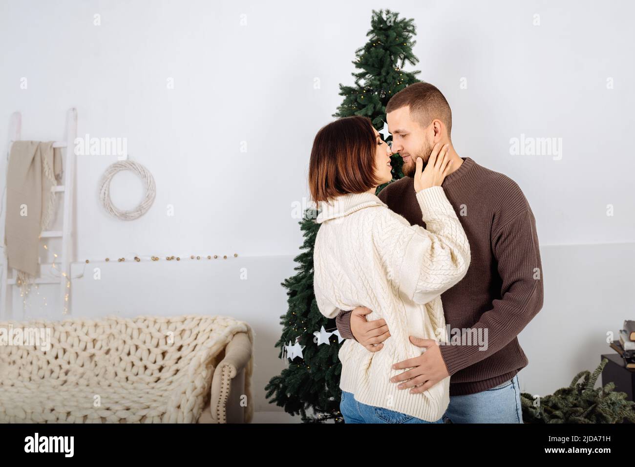 Beau jeune élégant couple, homme et femme, en pull blanc et brun dans les bras de l'autre sur le fond de l'arbre de Noël célébrer Noël à la maison. Minimalisme scandinave. Banque D'Images
