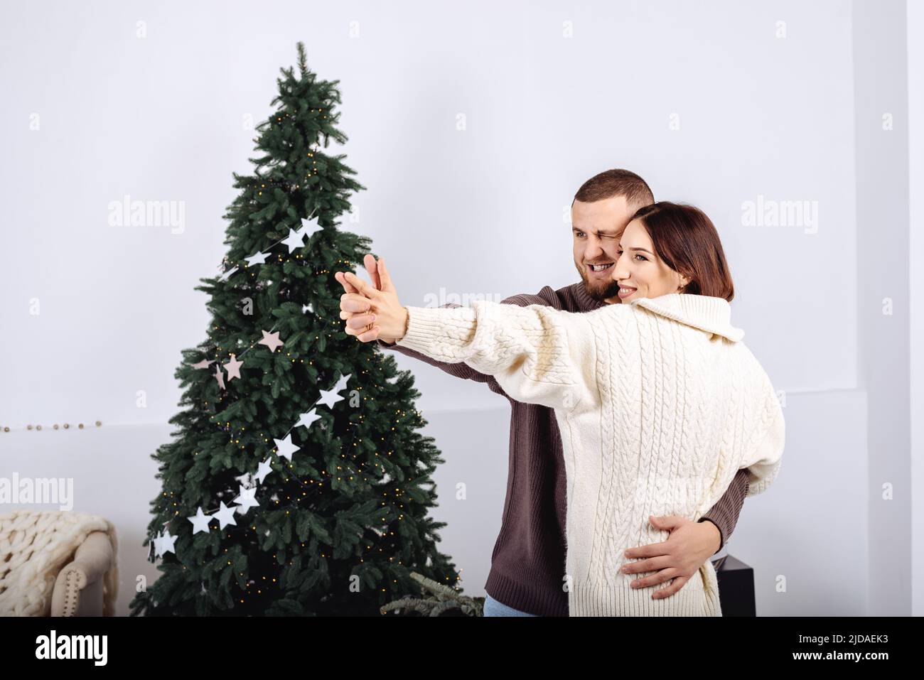 Joyeux jeune couple élégant, homme et femme dansant, dans des chandails tricotés blanc et brun dans les bras de l'autre sur fond d'arbre de Noël célébrant Noël à la maison. Minimalisme scandinave. Banque D'Images