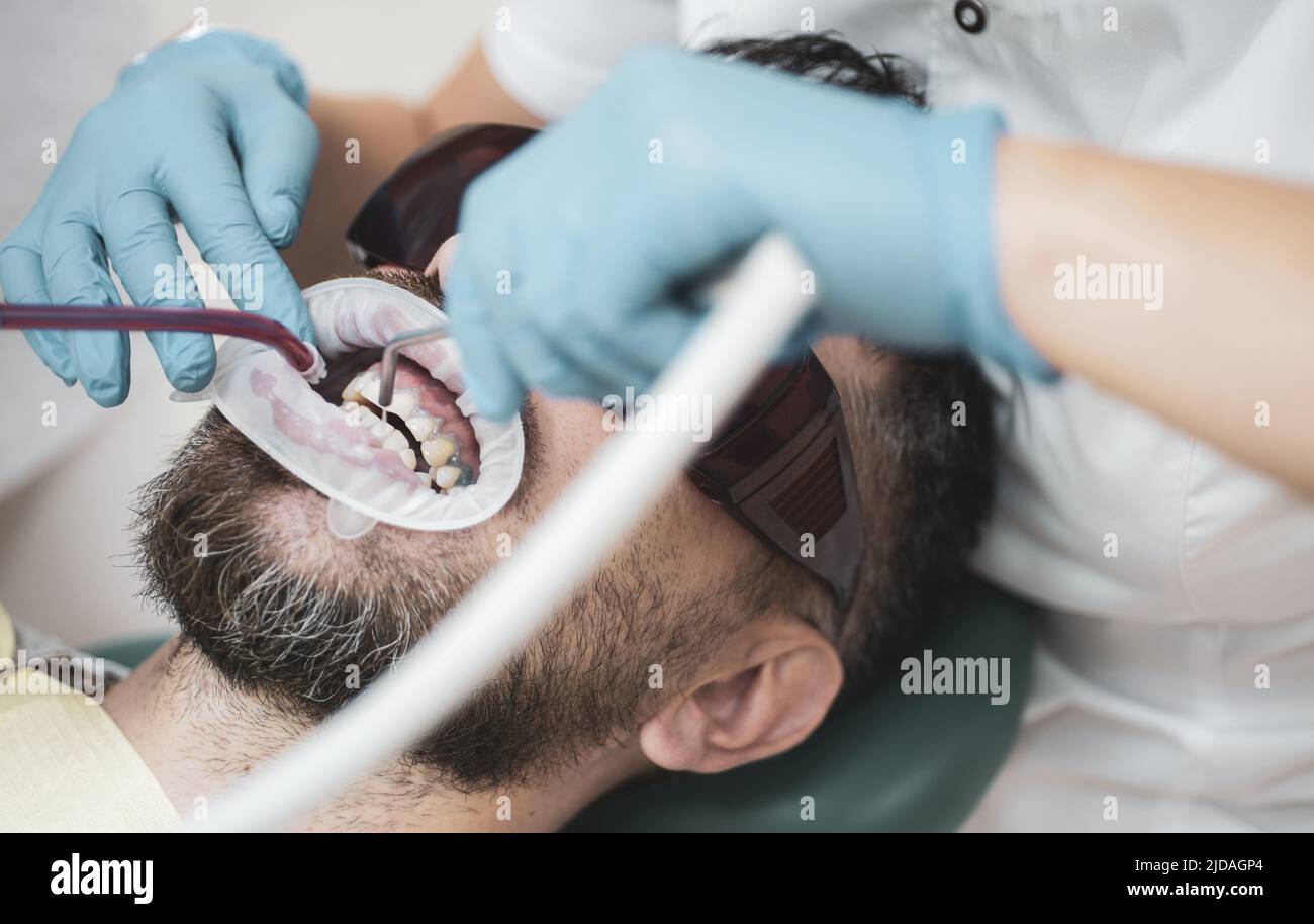 Jeune homme choisissant la couleur des dents chez le dentiste. Dentiste examinant les dents du patient en clinique. Jeune homme souriant après visite chez le dentiste sur vif flou Banque D'Images