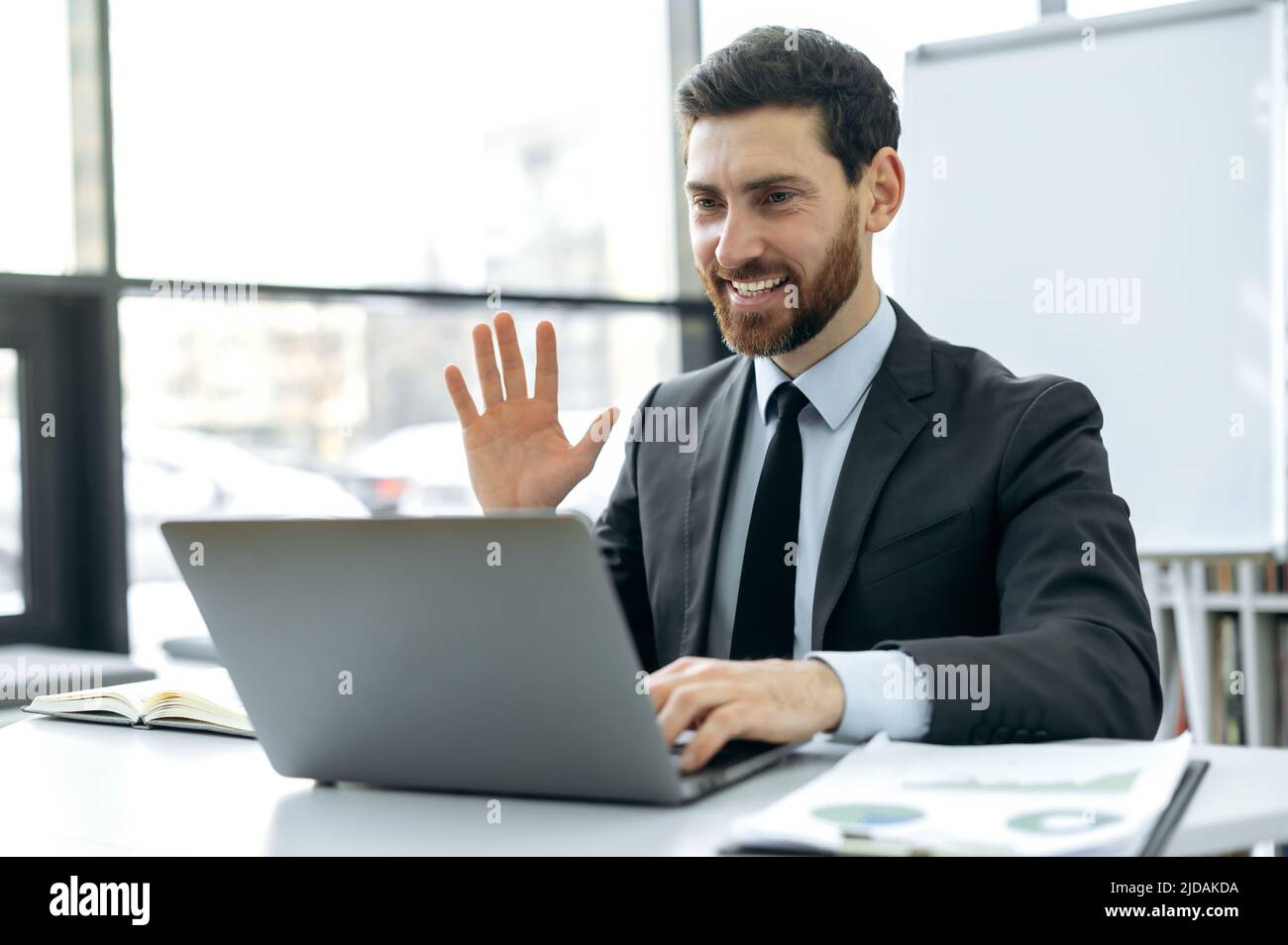 Homme caucasien positif avec une barbe, analyste financier, pdg de la société, assis dans un bureau moderne devant un écran d'ordinateur portable, ayant une réunion vidéo avec des collègues d'affaires, les saluant, souriant Banque D'Images