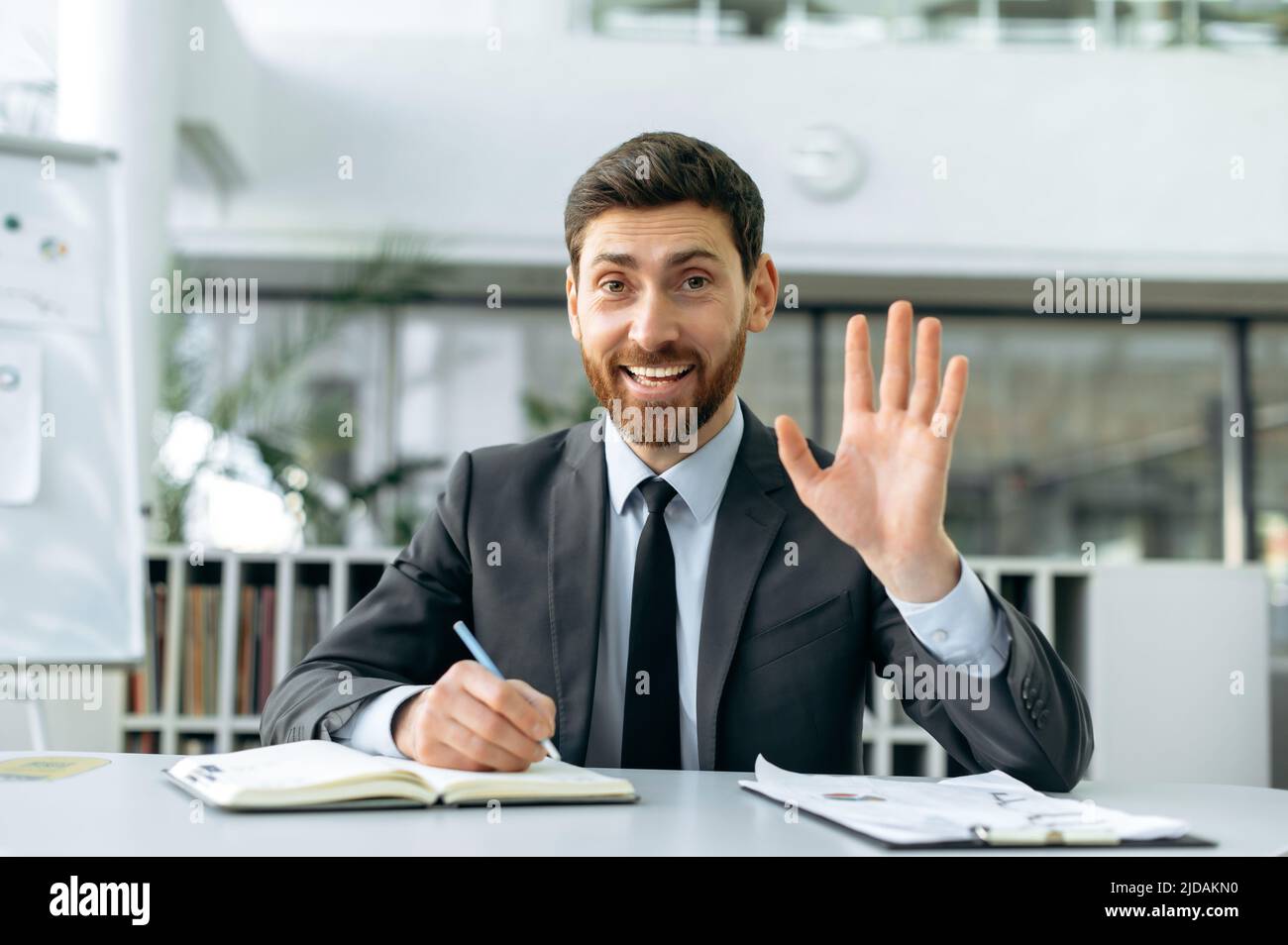 Ambitieux homme d'affaires caucasien positif, directeur d'entreprise, superviseur, assis dans un bureau moderne, dans des vêtements d'affaires, tenant une réunion vidéo en ligne avec des collègues, en agitant sa main dans le salut, sourires Banque D'Images
