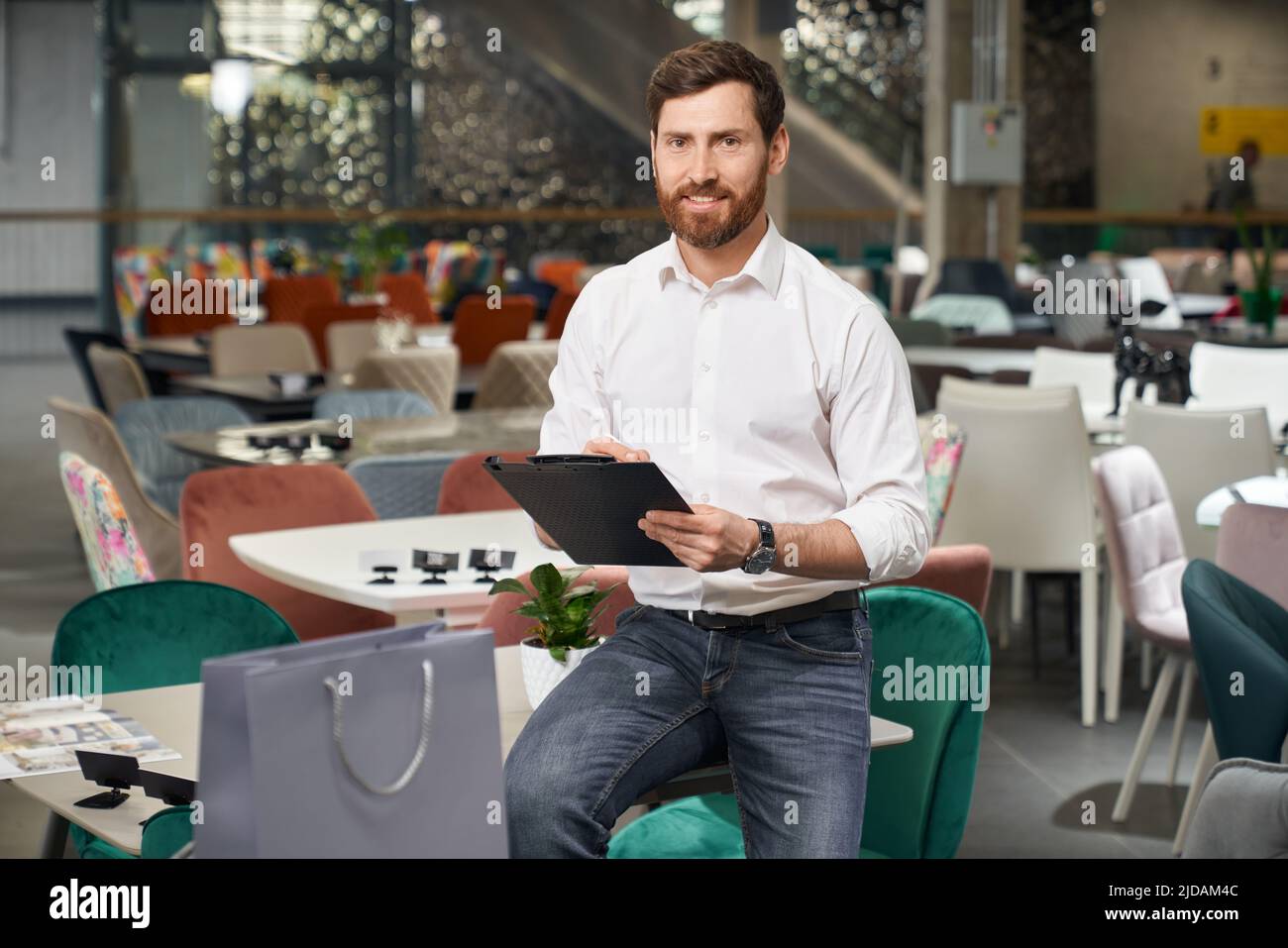 Vue de face d'un garçon à la mode assis sur une table, d'un dossier de conservation, de l'écriture. Beau concepteur, vendeur, architecte conception, dossier de maintien, regardant la caméra. Concept de conception. Banque D'Images Vue de face d'un garçon à la mode assis sur une table, d'un dossier de conservation, de l'écriture. Beau concepteur, vendeur, architecte conception, dossier de maintien, regardant la caméra. Concept de conception. Banque D'Images