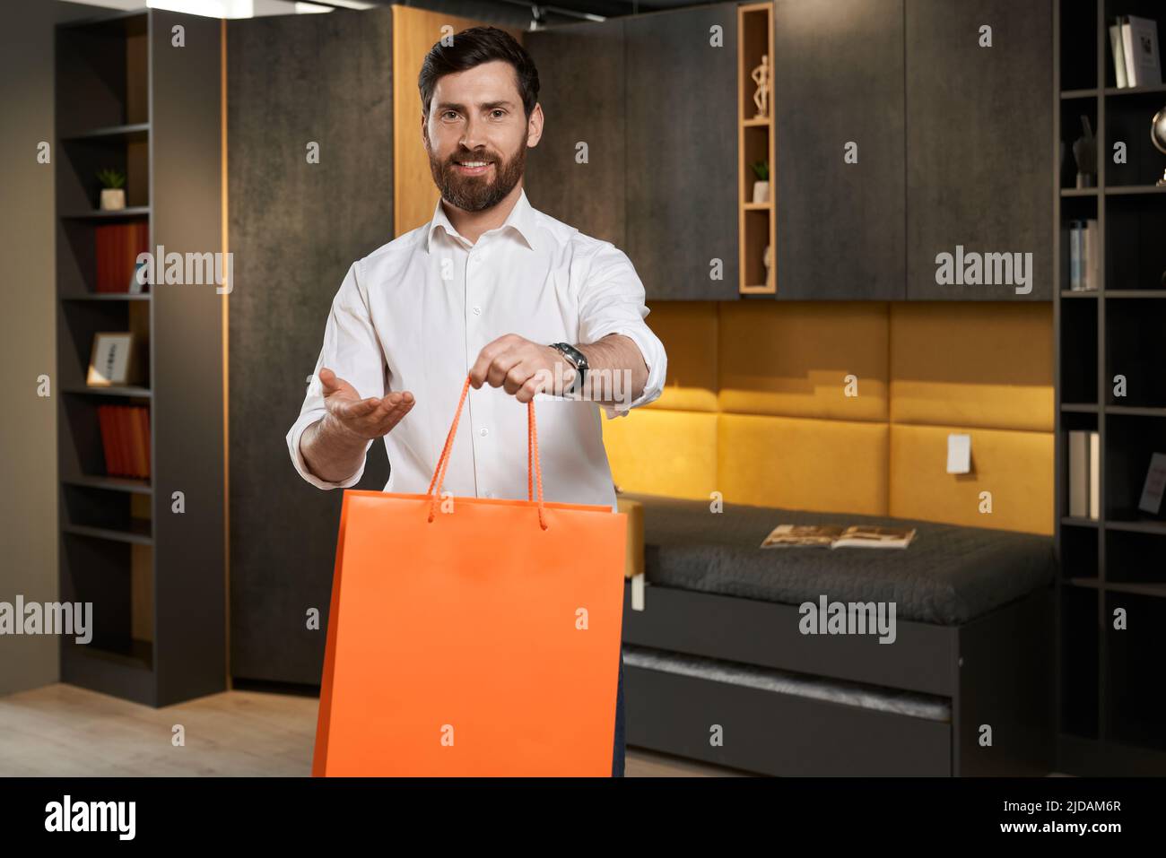 Vue avant de la belle position du consommateur, tenue, montrant le sac de don dans le magasin de meubles. Brunette homme souriant, regardant l'appareil photo, heureux, heureux, satisfait. Concept de shopping. Banque D'Images Vue avant de la belle position du consommateur, tenue, montrant le sac de don dans le magasin de meubles. Brunette homme souriant, regardant l'appareil photo, heureux, heureux, satisfait. Concept de shopping. Banque D'Images