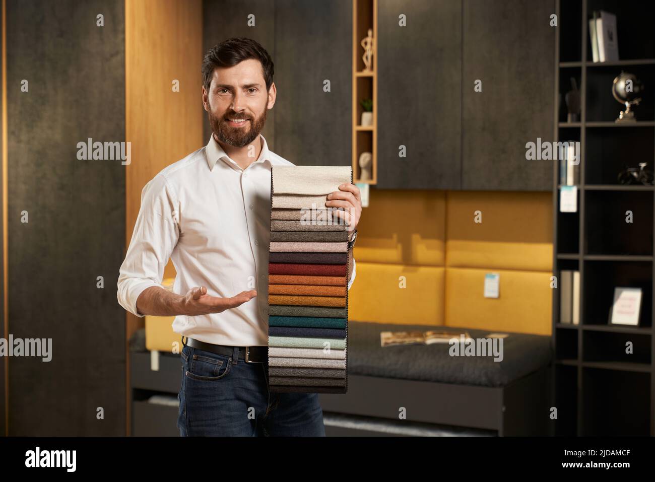 Vue avant du vendeur heureux debout, montrant, représentant la palette de tissus pour les meubles. Beau homme avec barbe regardant l'appareil photo, souriant. Concept d'achat de meubles. Banque D'Images Vue avant du vendeur heureux debout, montrant, représentant la palette de tissus pour les meubles. Beau homme avec barbe regardant l'appareil photo, souriant. Concept d'achat de meubles. Banque D'Images
