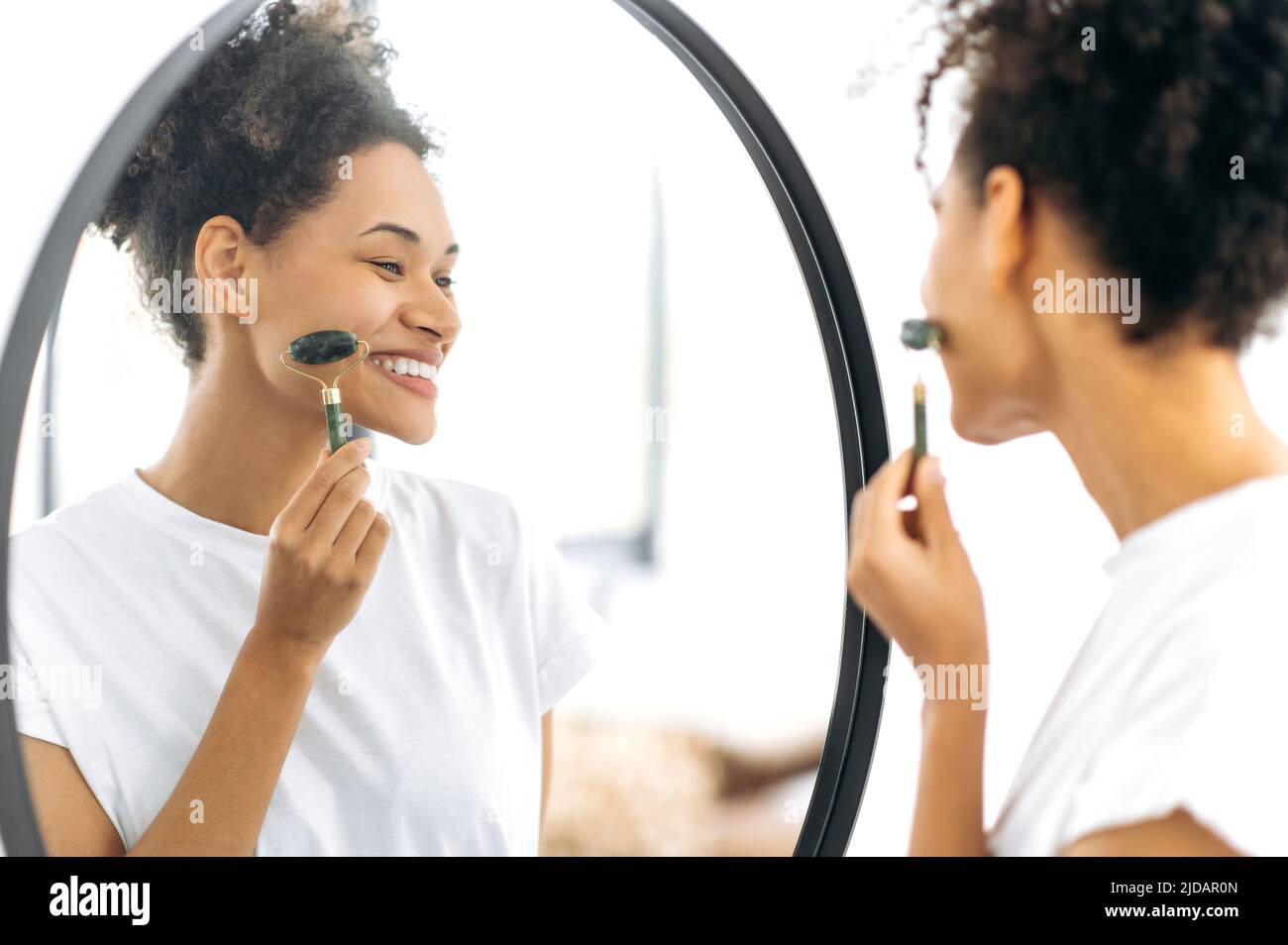 Soin du visage, prévention des rides. Belle jeune femme afro-américaine aux cheveux bouclés, faisant des procédures de soin de la peau du visage, debout devant un miroir à la maison, en utilisant un rouleau de jade, souriant heureux Banque D'Images