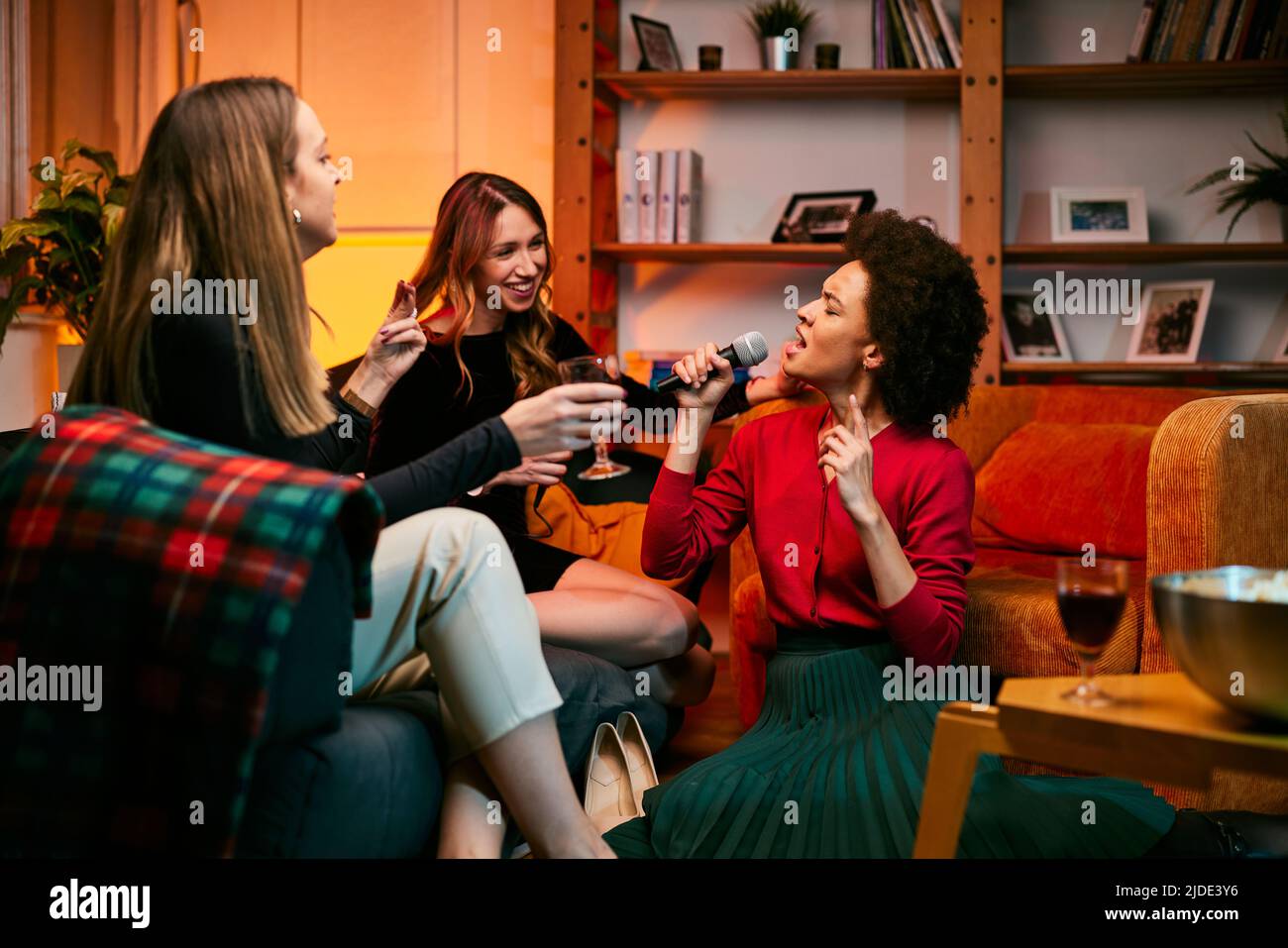 Un groupe multiculturel d'amis féminins chante karaoké tout en buvant du vin dans le salon. Banque D'Images