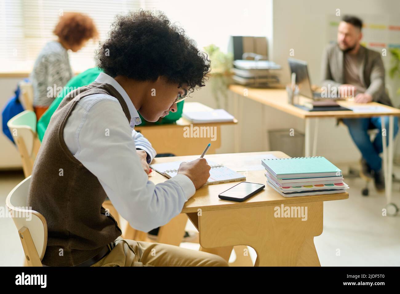 Élève de haute école en vêtements décontractés intelligents faire des notes en copybook à la leçon tout en effectuant la tâche individuelle contre les camarades de classe et l'enseignant Banque D'Images