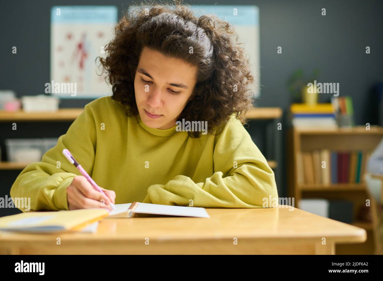 Adolescent de sexe masculin dans un pull-over jaune prenant des notes dans son copybook tout en étant assis au bureau à la leçon et en effectuant une tâche individuelle Banque D'Images