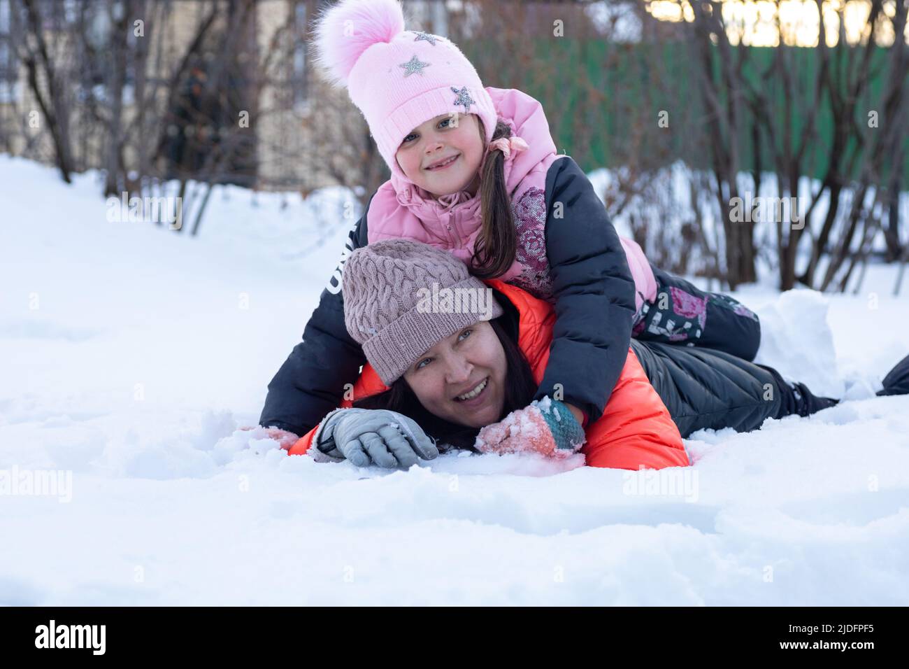 Petite fille heureuse de race blanche avec mère couché sur la neige ensemble souriant dans le parc le soir avec des arbres en arrière-plan. Les parents passent du temps avec Banque D'Images