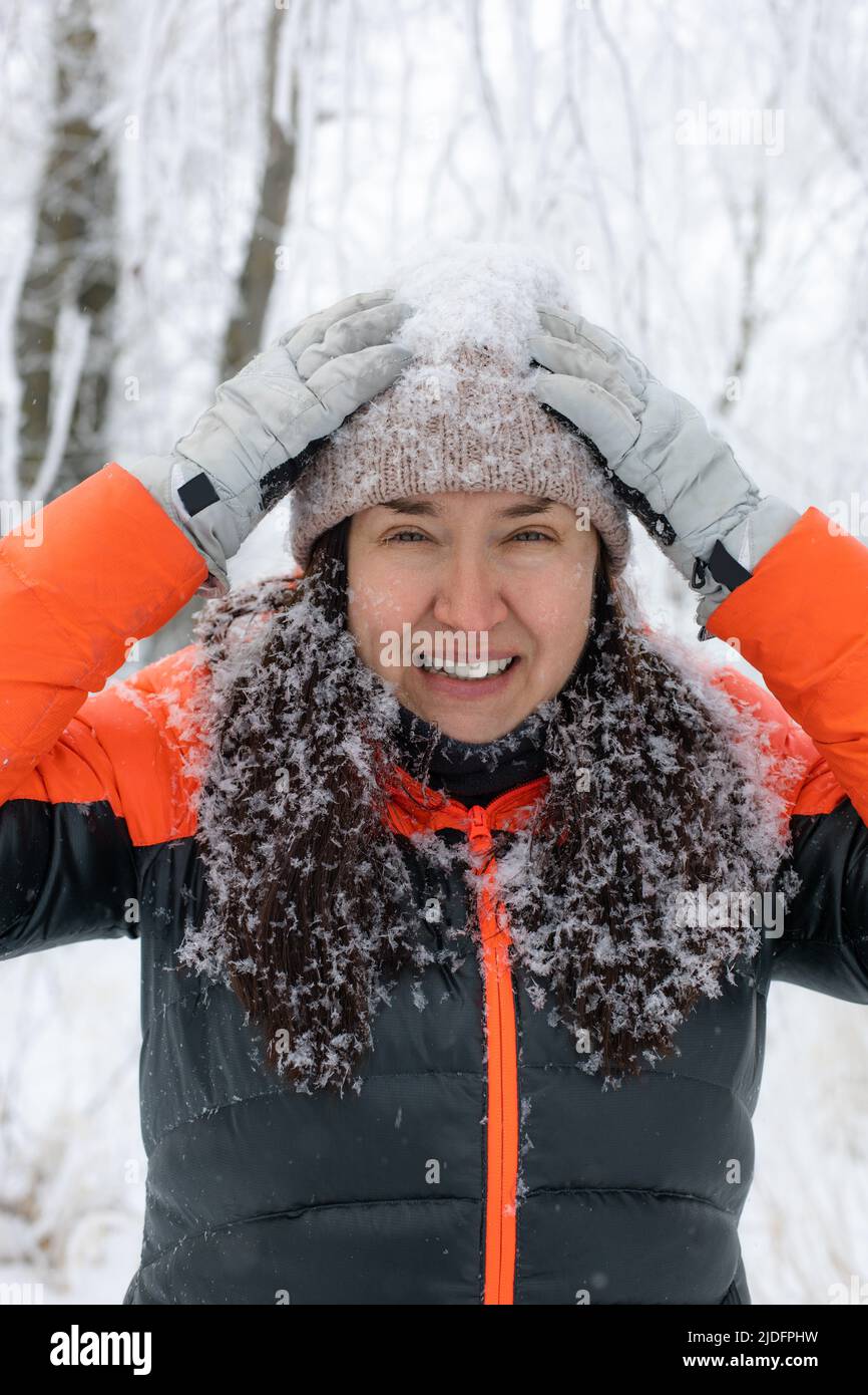 Portrait d'une femme souriante d'âge moyen regardant l'appareil photo touchant un chapeau avec les mains dans des gants habillés de vêtements chauds d'hiver avec les cheveux, chapeau recouvert de Banque D'Images