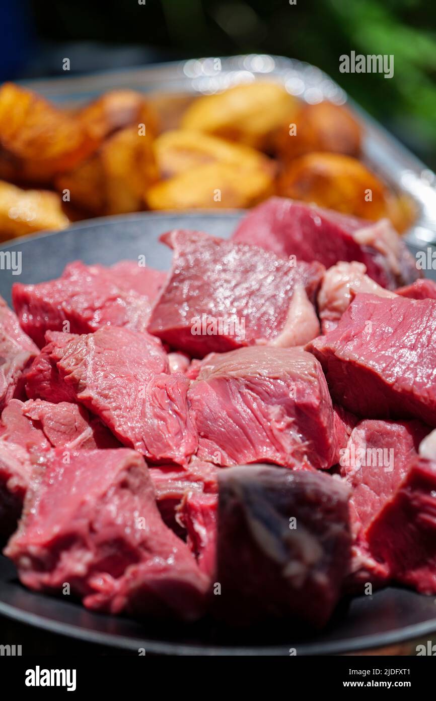 Gros plan de morceaux de viande de bœuf crus coupés en dés sur une plaque en aluminium et de pommes de terre rôties en arrière-plan. Banque D'Images