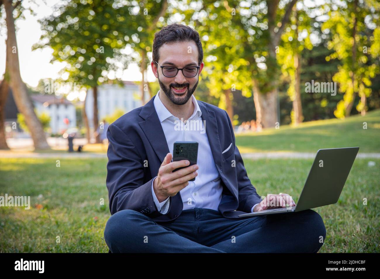 Homme d'affaires assis dans un pré utilise son ordinateur portable et regarde l'écran de son smartphone Banque D'Images