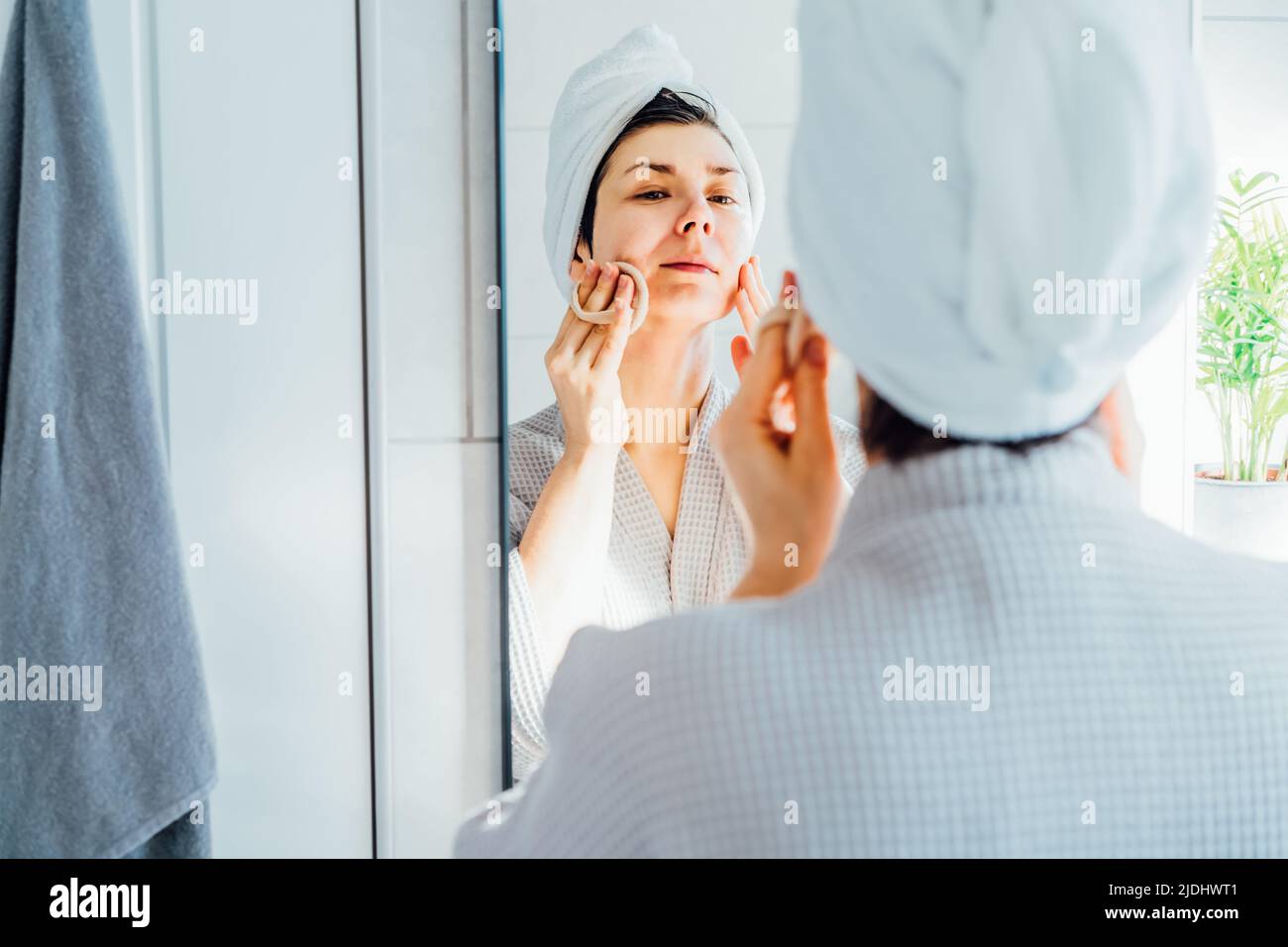 Femme en peignoir avec une serviette sur la tête regardant dans le miroir, exfoliant la peau du visage avec l'éponge loofah, enlever le maquillage ou faire la routine de nettoyage, la peau Banque D'Images Femme en peignoir avec une serviette sur la tête regardant dans le miroir, exfoliant la peau du visage avec l'éponge loofah, enlever le maquillage ou faire la routine de nettoyage, la peau Banque D'Images