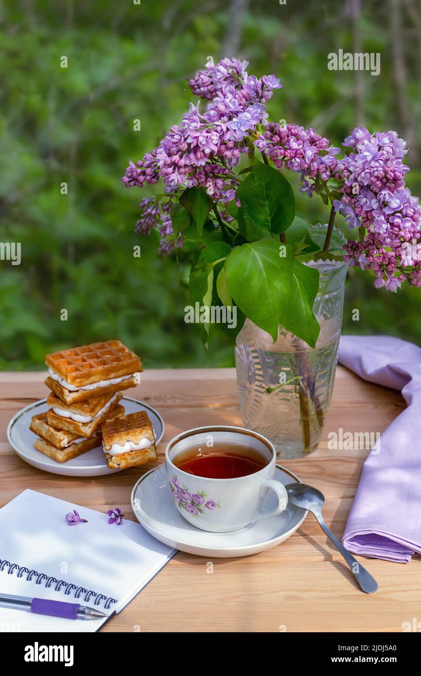 Une fête de thé avec des gaufres viennoises et une branche de lilas et un carnet vide ouvert pour écrire sur une table en bois sur le fond d'un jardin Banque D'Images